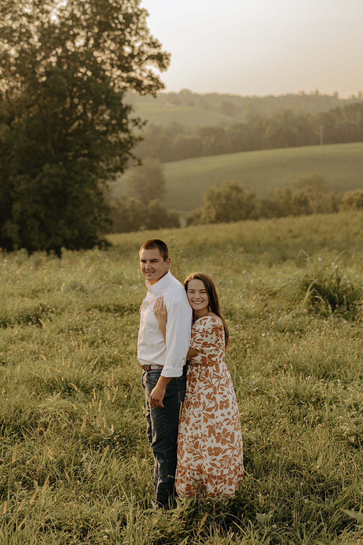 A couple stands together in a grassy field during golden hour with rolling hills in the background.