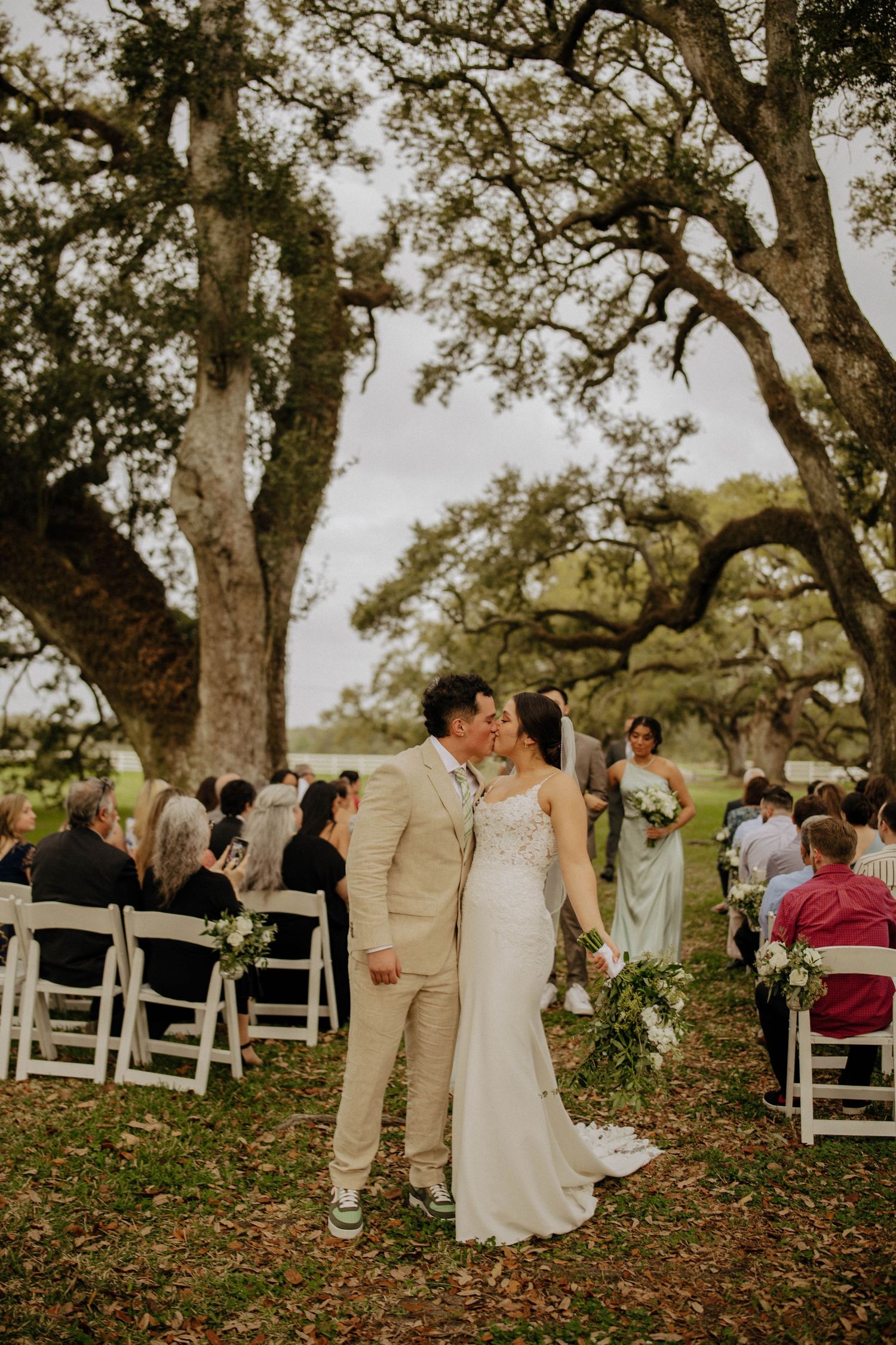 Outdoor wedding ceremony under majestic oak trees with scattered white chairs on autumn ground.