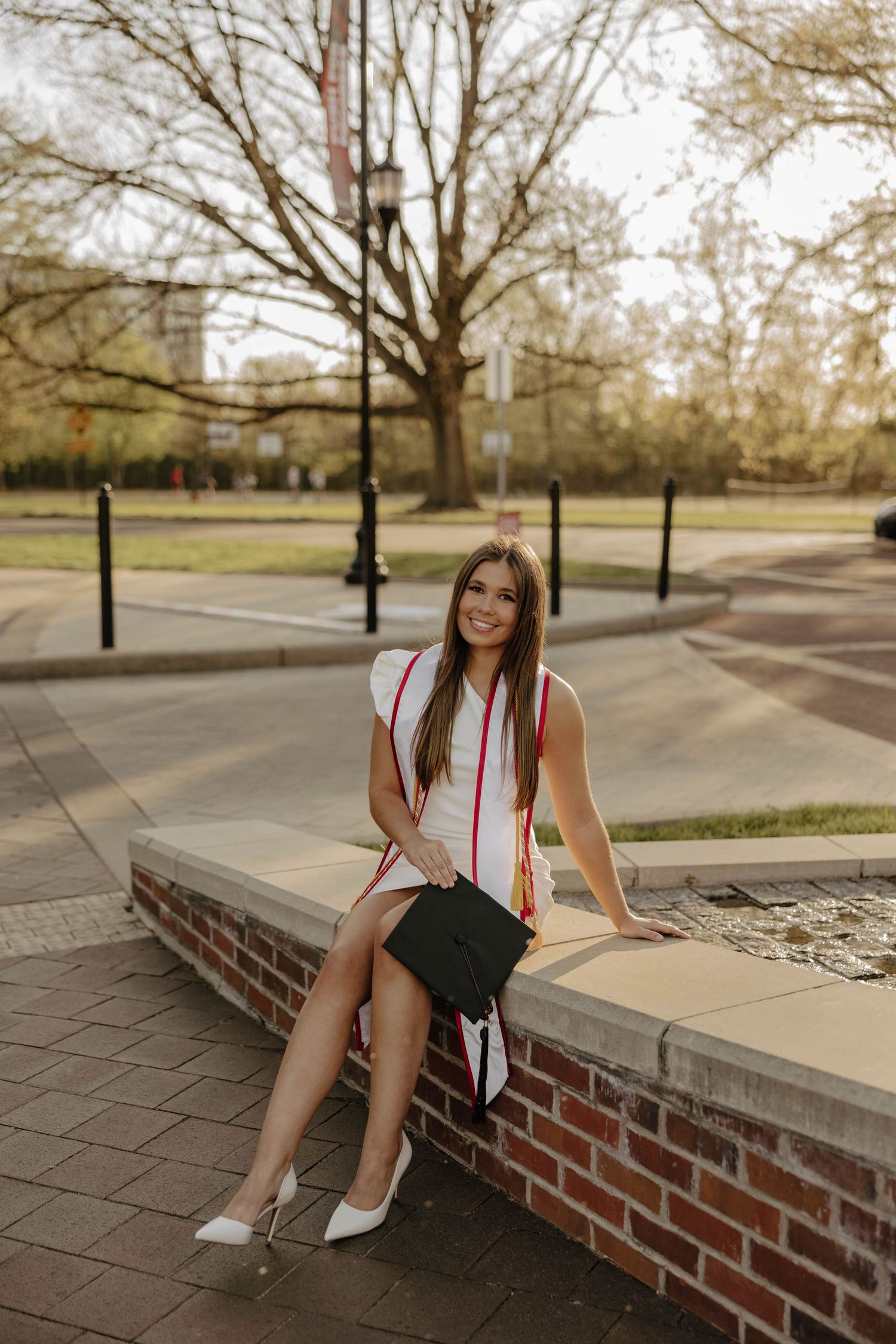 Graduate in white dress and red stoles sitting on brick wall during outdoor photo shoot.