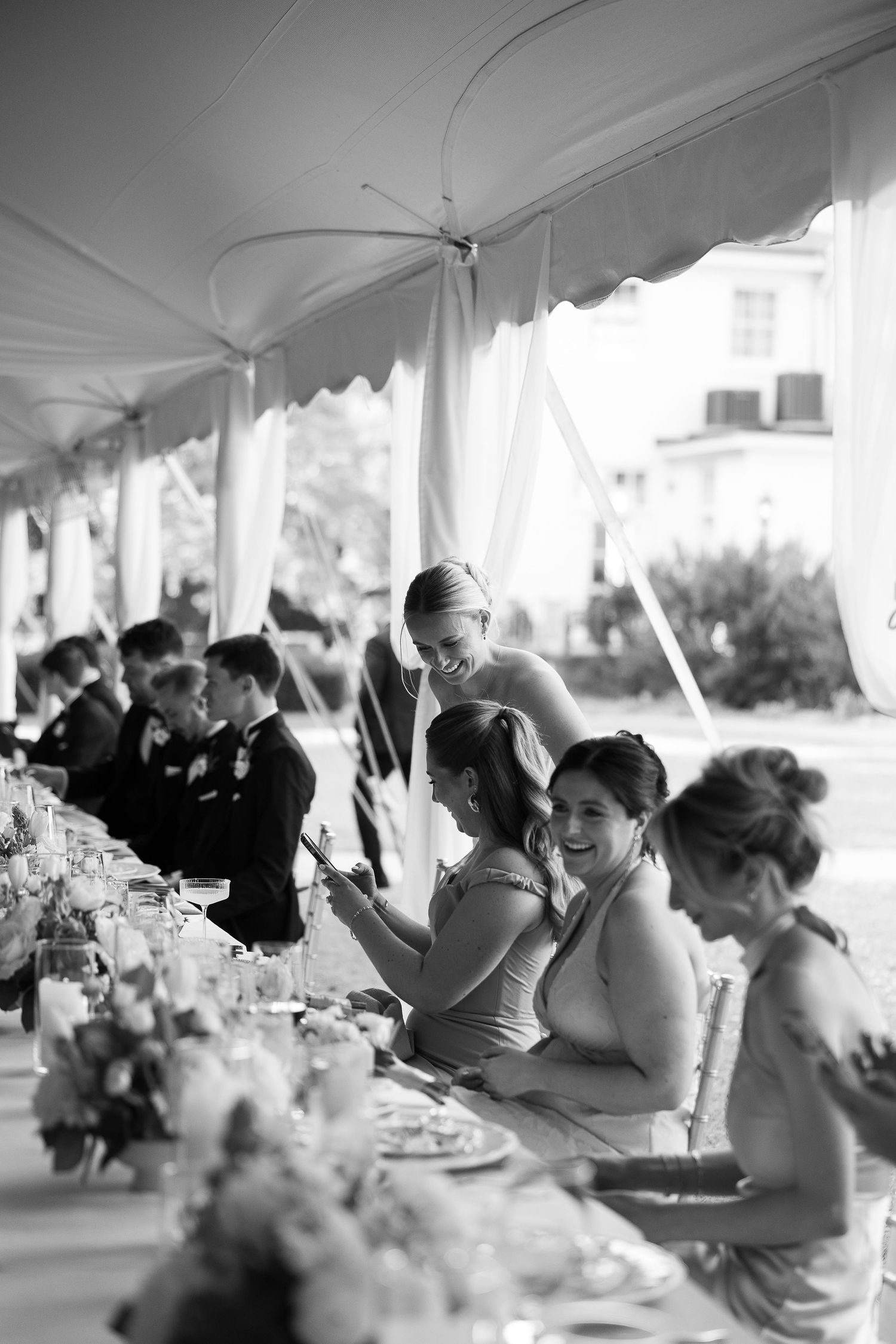 Black and white photo of bridesmaids laughing together at elegantly set outdoor wedding reception table.