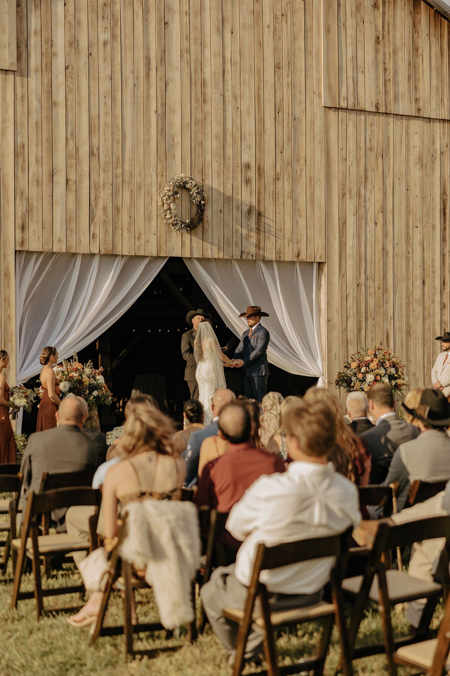 Rustic barn wedding ceremony with white drapes and floral decorations as guests watch from wooden chairs.