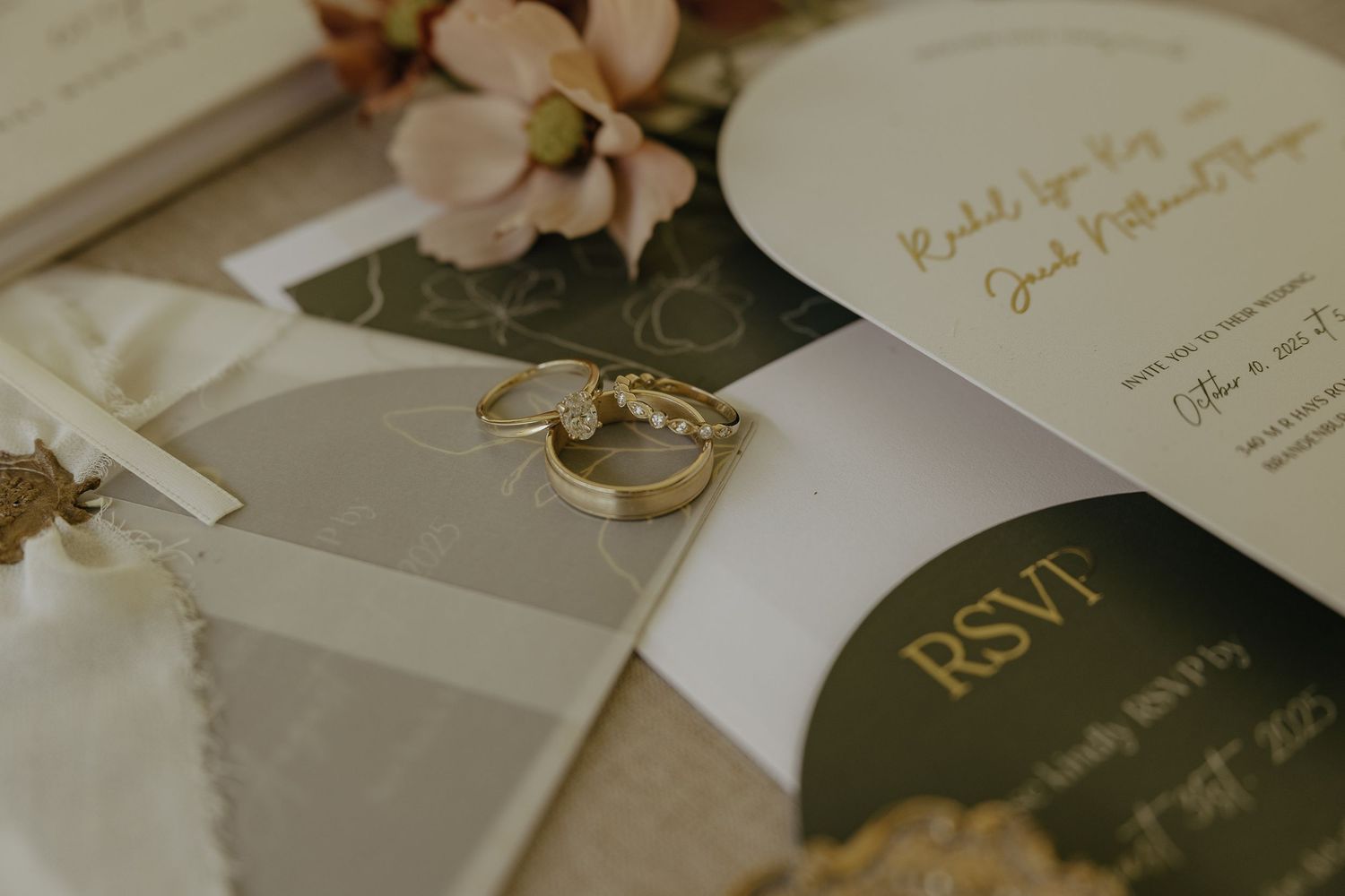 Wedding rings rest on elegant invitation cards with blush pink floral accents and gold calligraphy details.