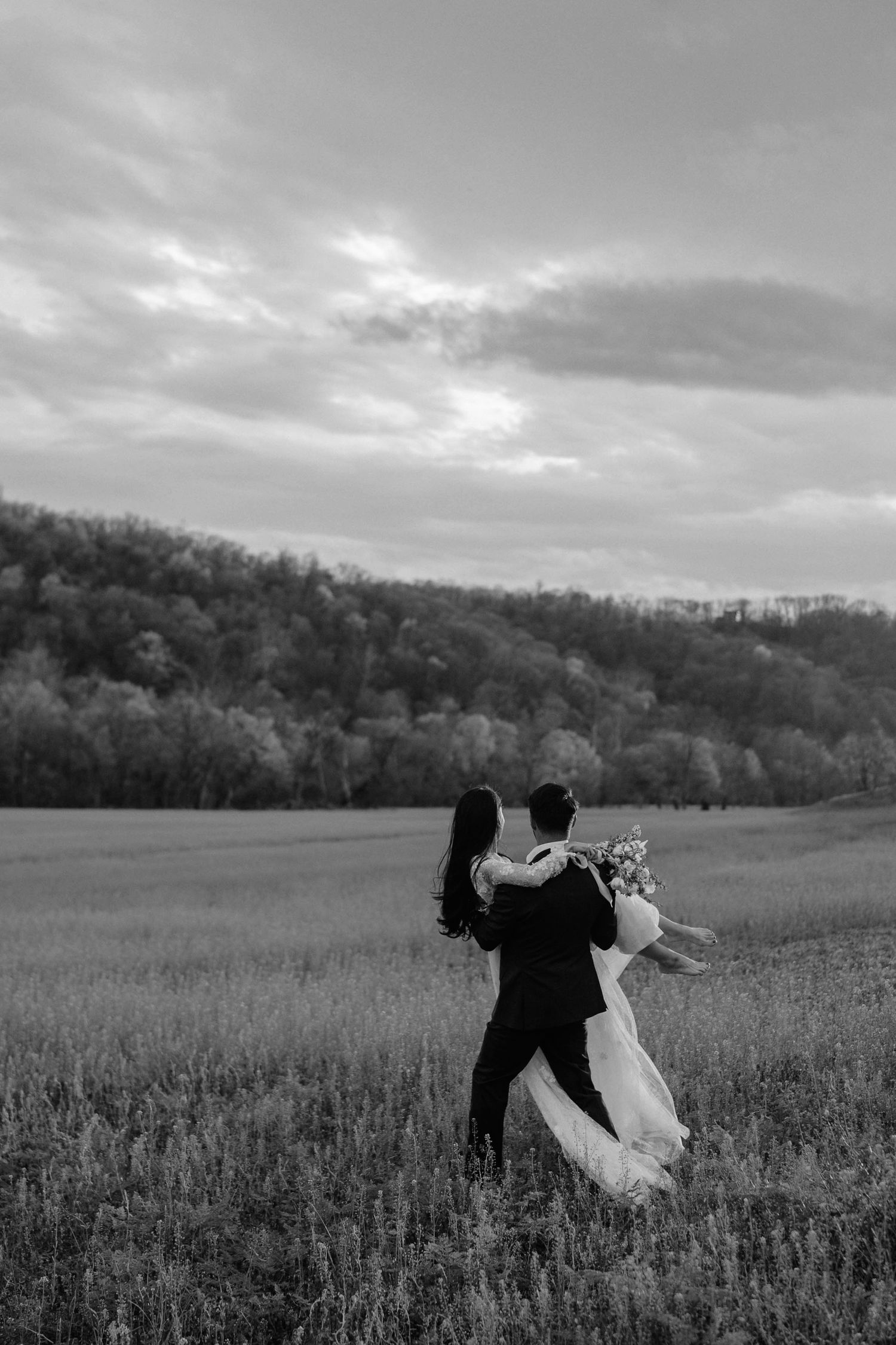 A couple dancing together in a scenic meadow with rolling hills and cloudy sky in black and white photography.