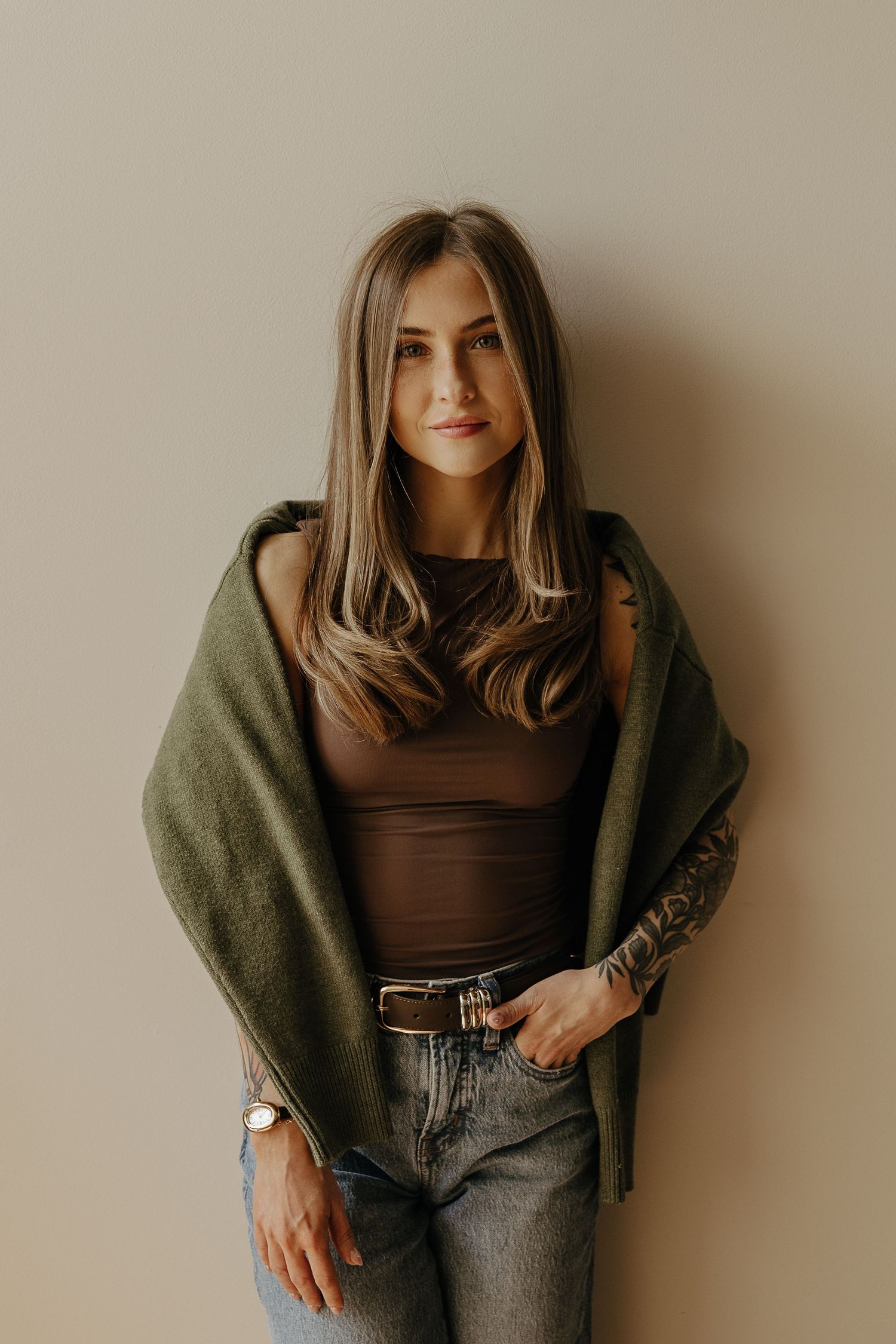 Young woman with long hair wearing a brown top, green shawl, jeans, and a watch, posing against a beige wall.