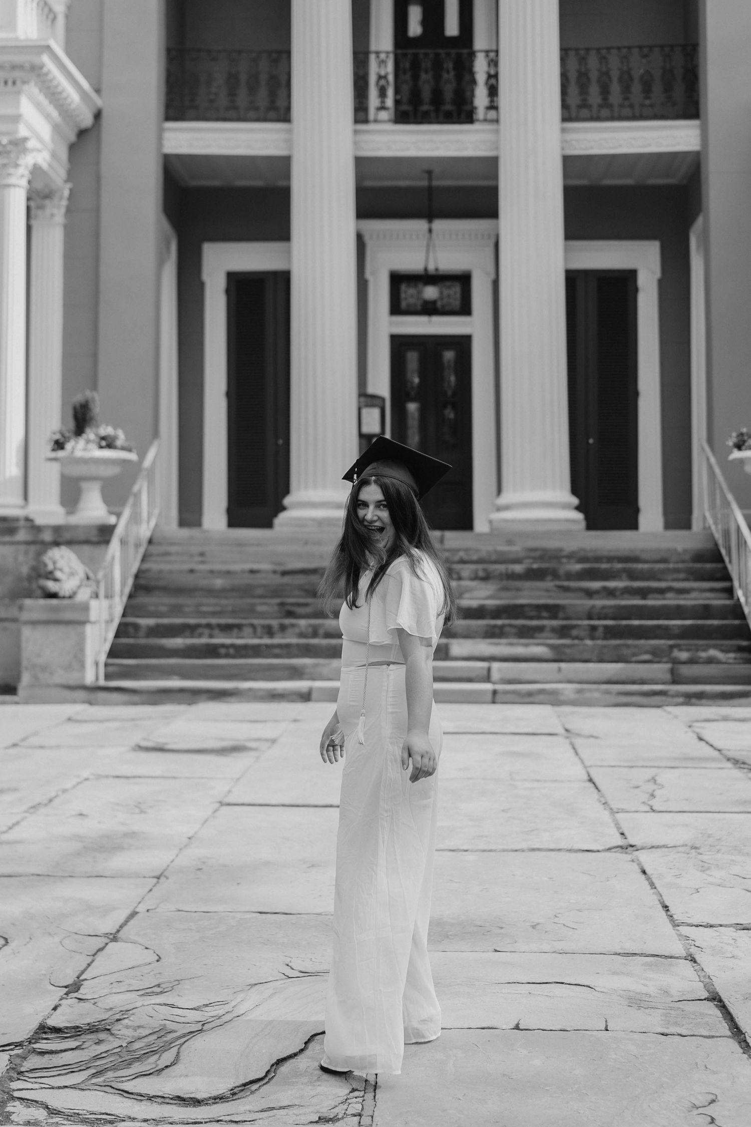 A graduate poses in front of a neoclassical building with white columns on a stone courtyard.