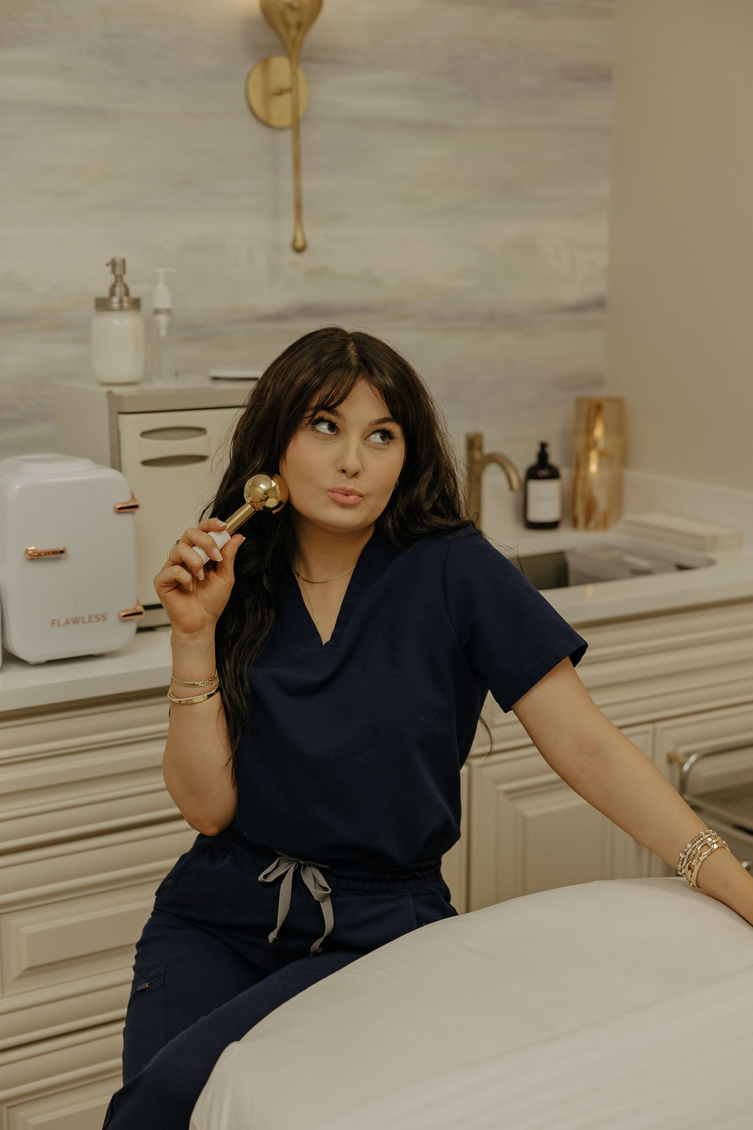 Medical professional in navy scrubs poses on treatment table in modern aesthetic clinic.