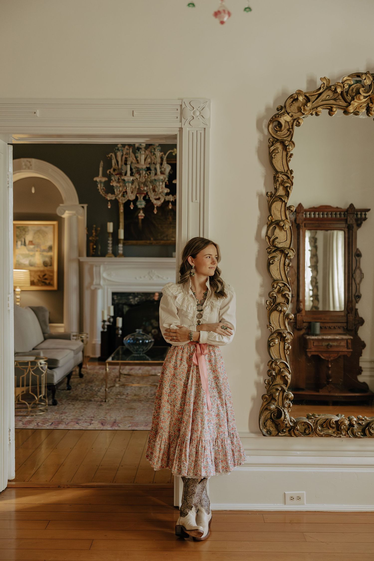 A pink floral dress is reflected in an ornate gold mirror in an elegant room with hardwood floors.