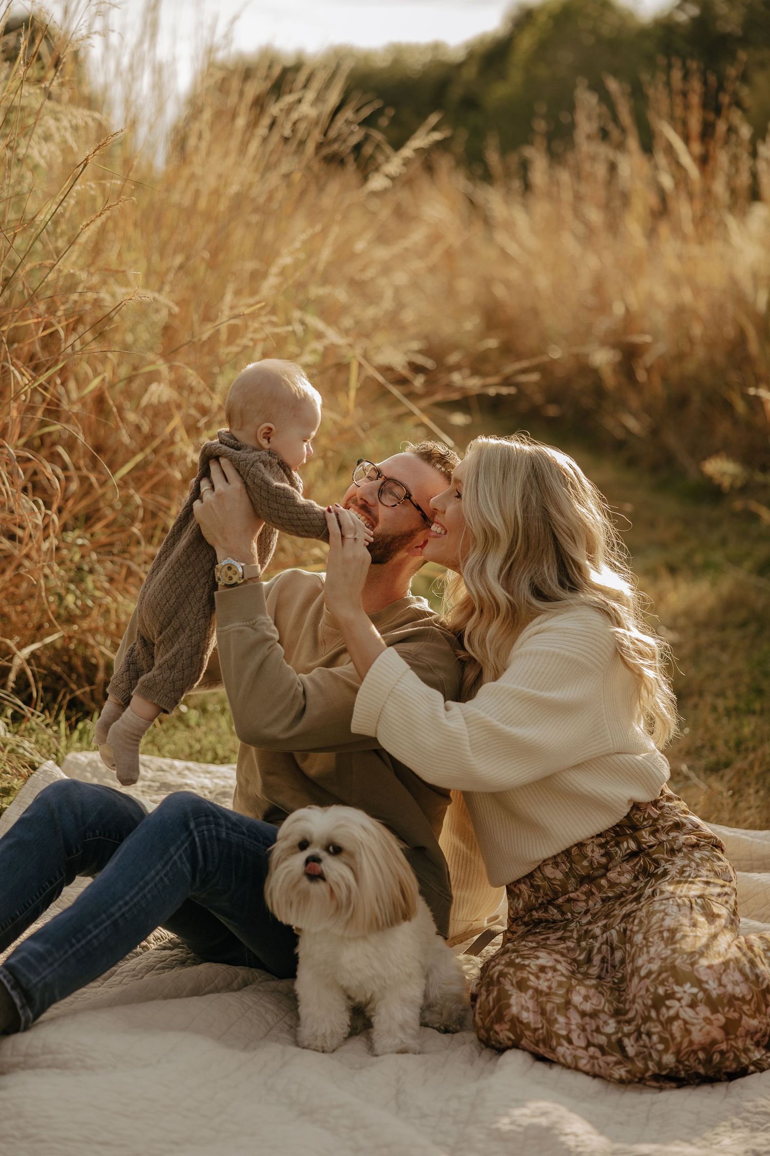 Playful family poses on blanket with white dog during sunset photoshoot.