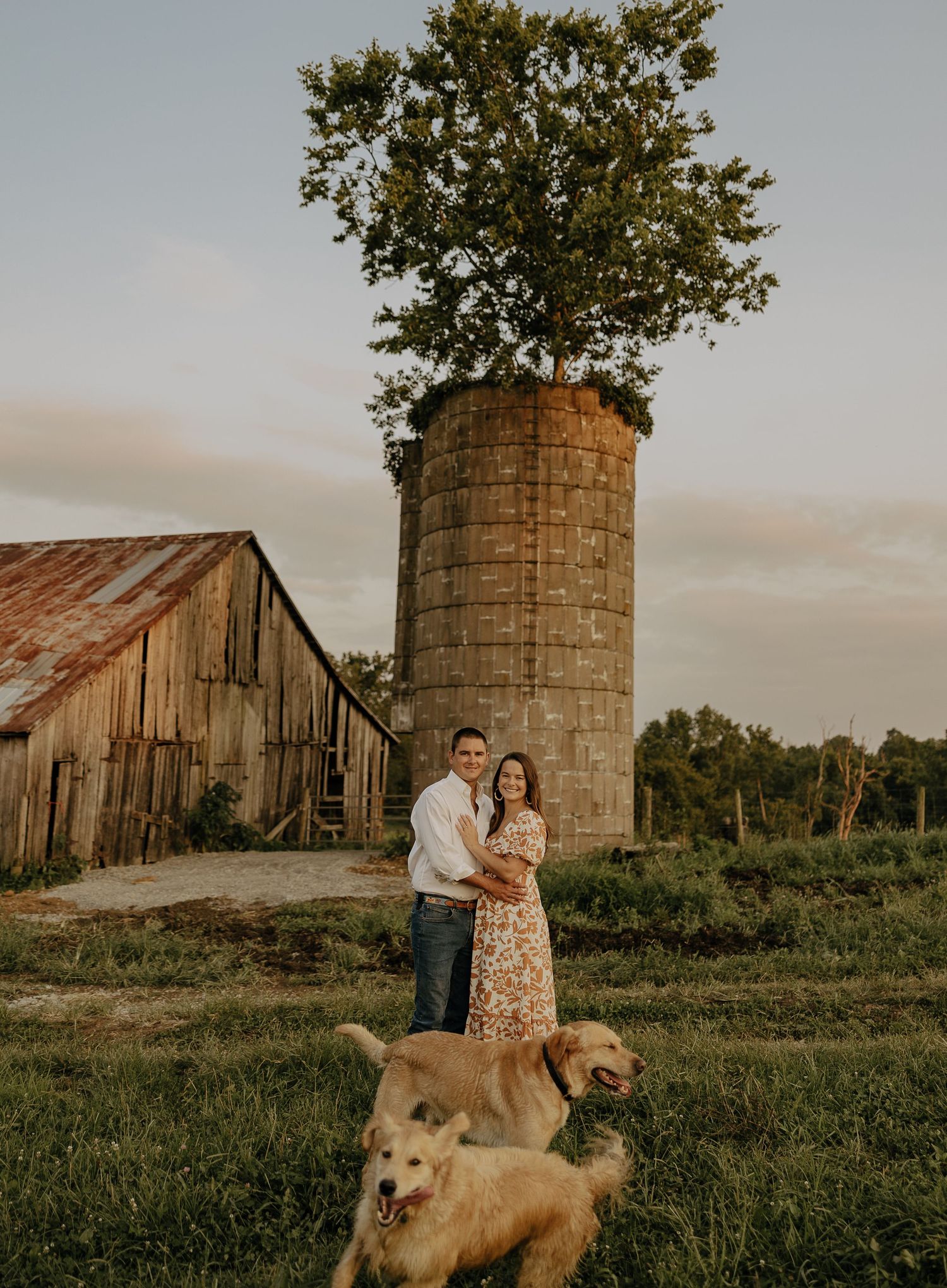 Couple poses with their golden retriever near rustic barn and silo at sunset.