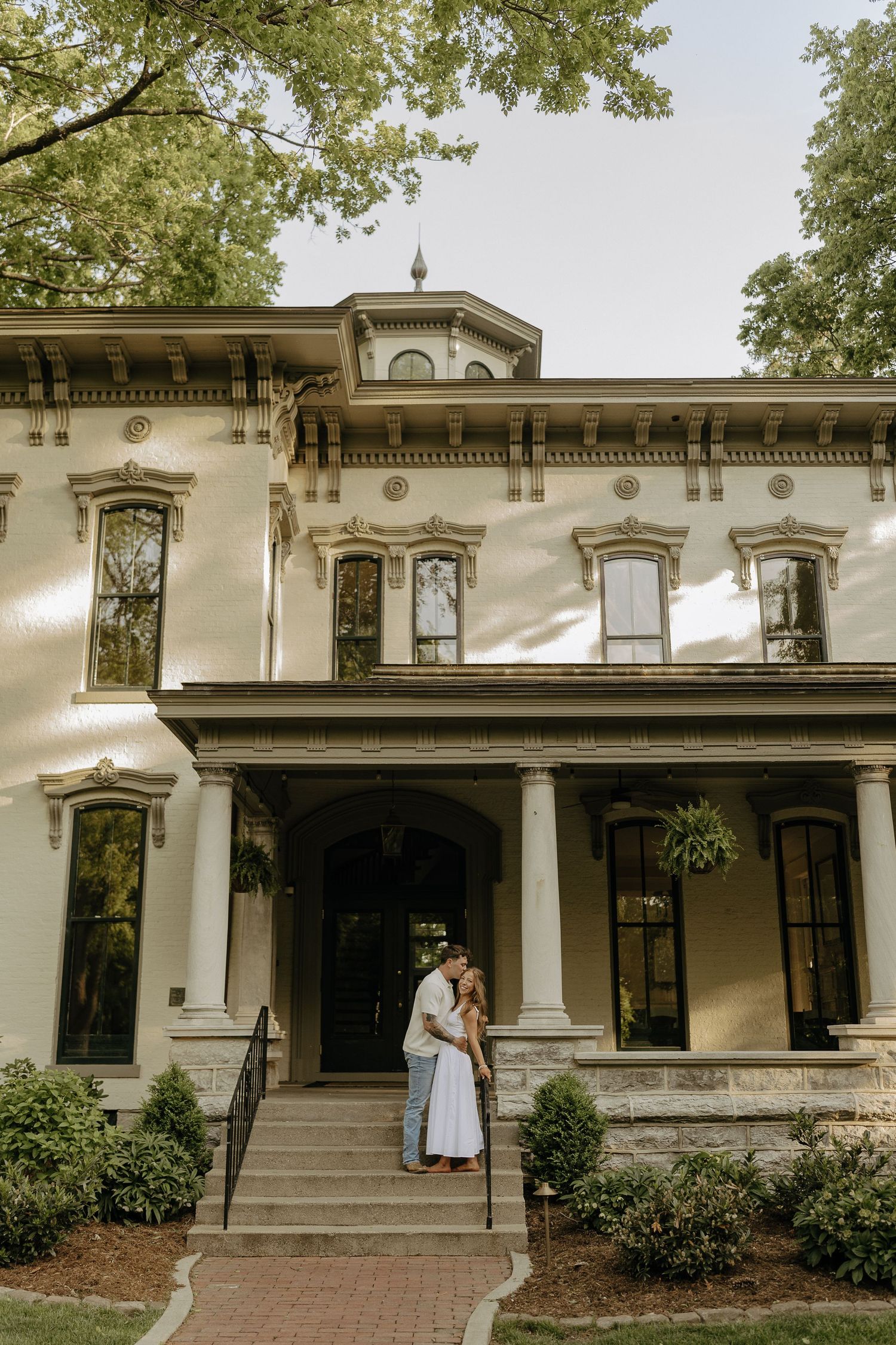 Couple sharing a romantic embrace on the steps of a historic Victorian mansion surrounded by lush greenery.