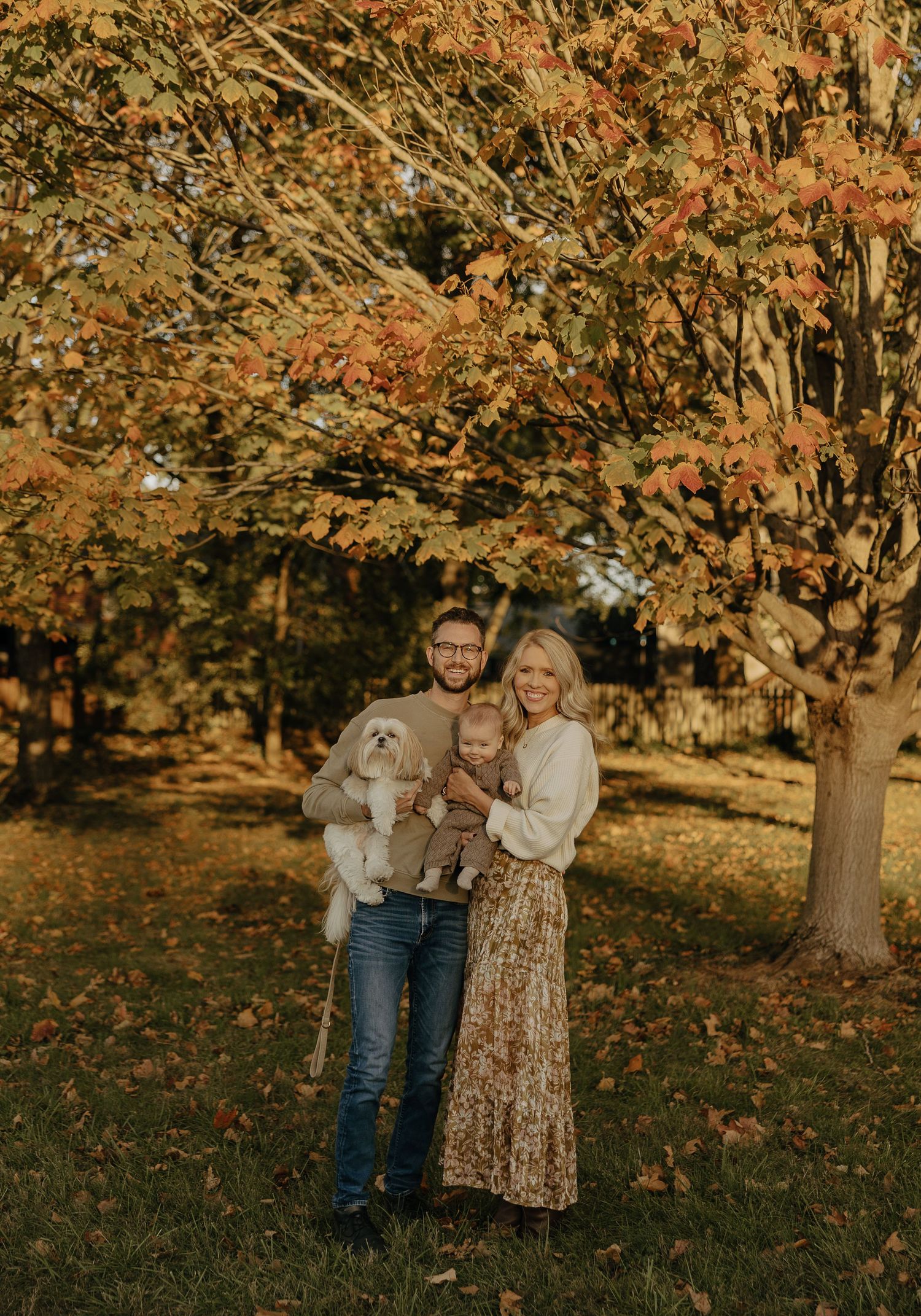 A couple poses with their white dog against autumn trees with orange leaves.