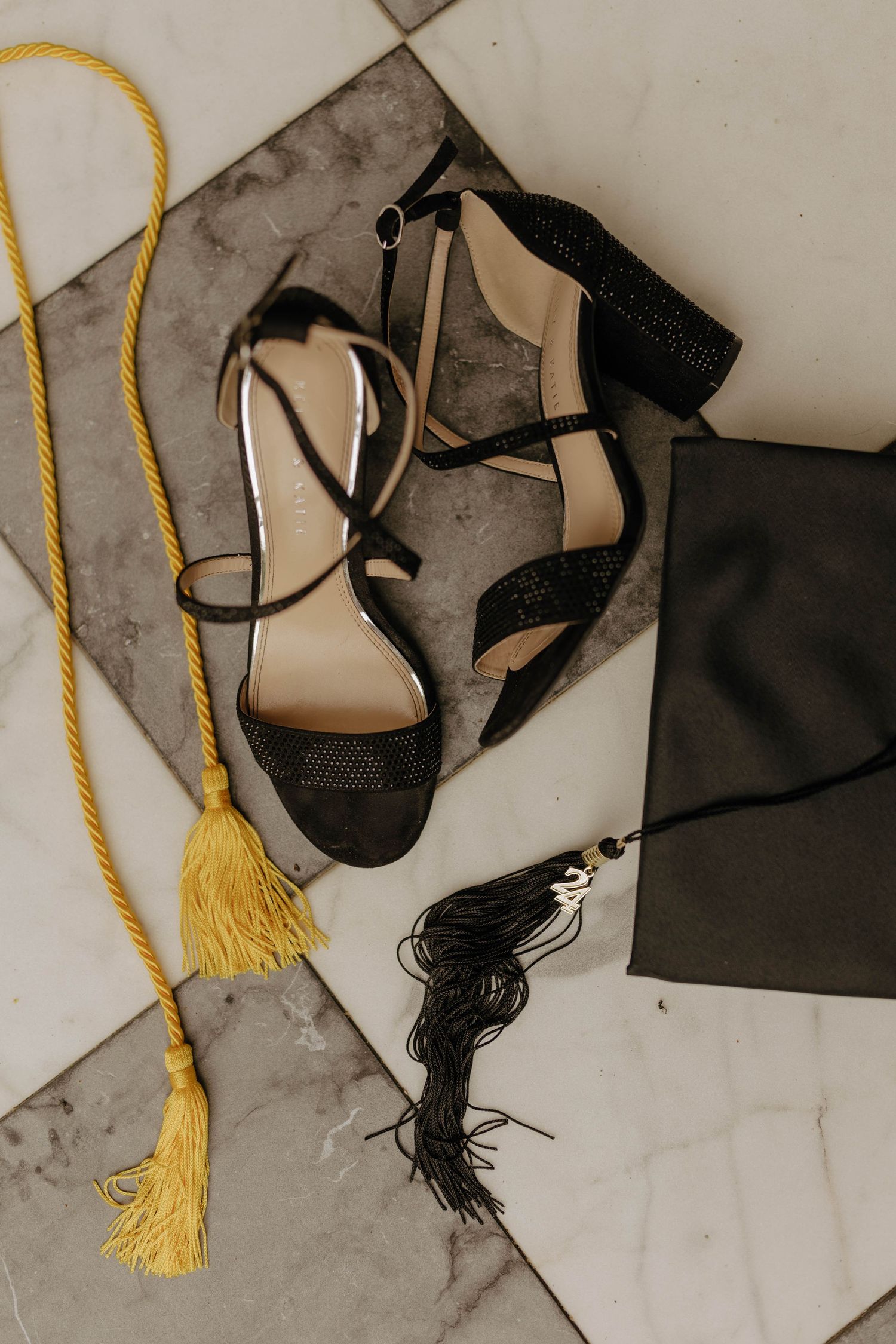 Black graduation cap and tassel arranged with formal shoes on marble surface.