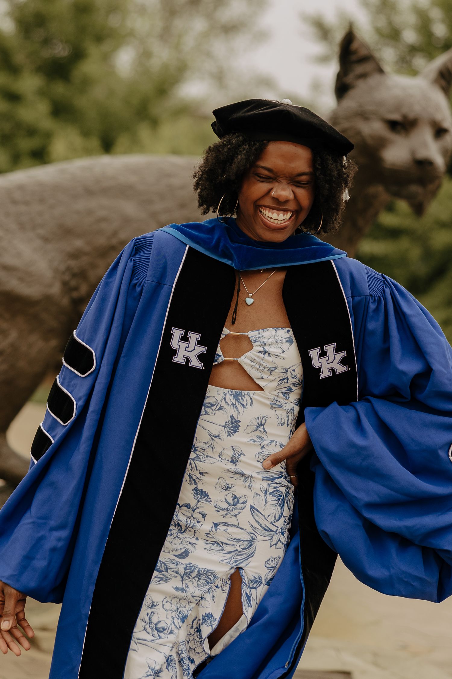 Graduate wearing blue and black academic regalia with University of Kentucky emblems poses joyfully outdoors.