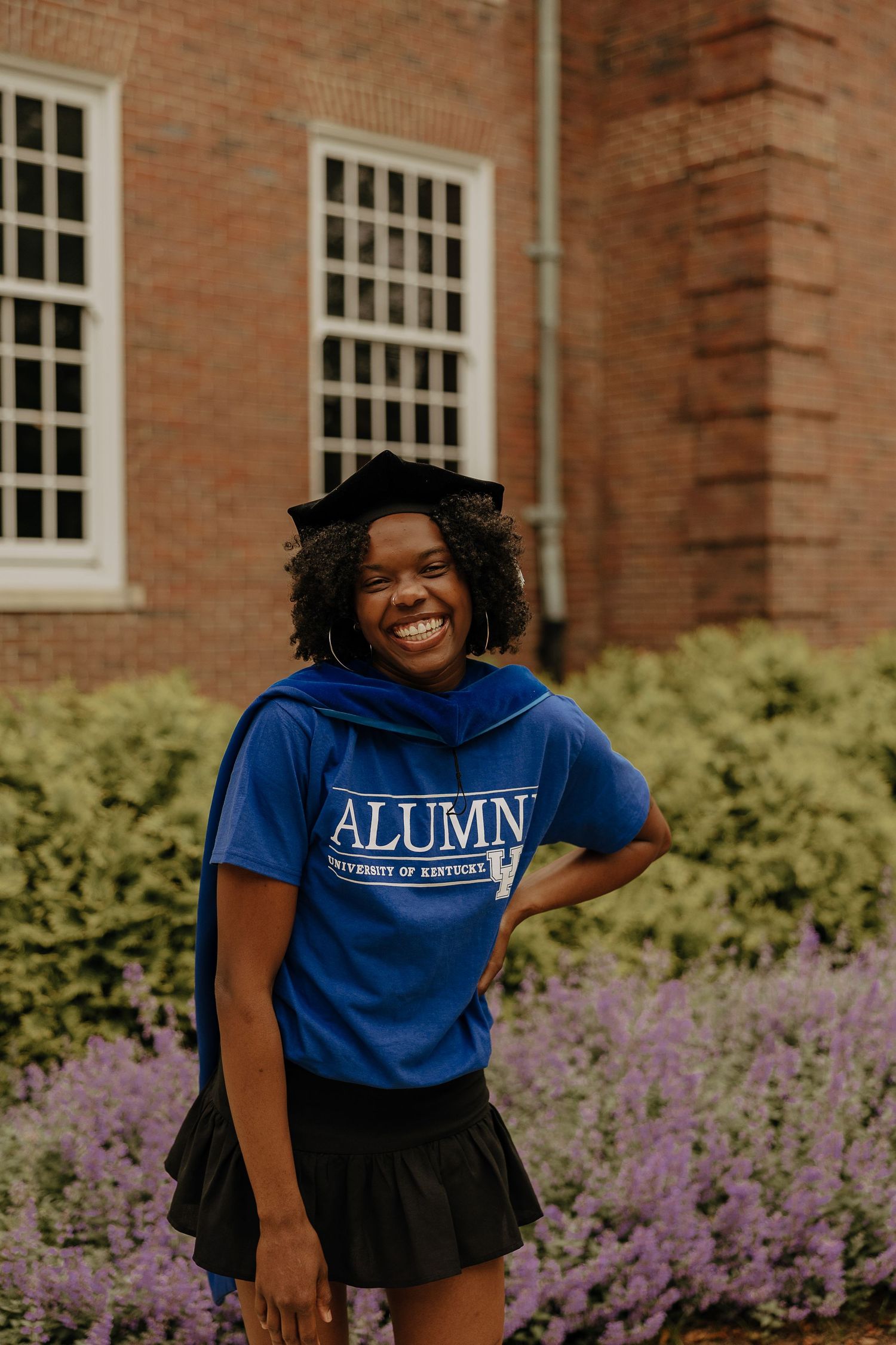 Purple flowers bloom in front of a brick building with an alumni wearing a blue sweatshirt.