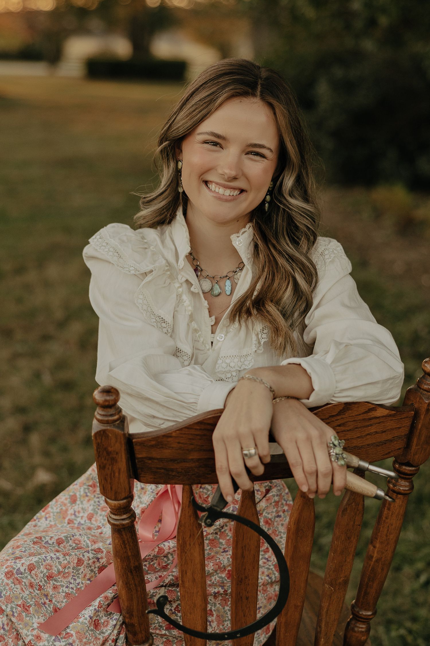 Portrait in warm lighting showcasing wavy hair and jewelry while seated in a wooden chair outdoors.