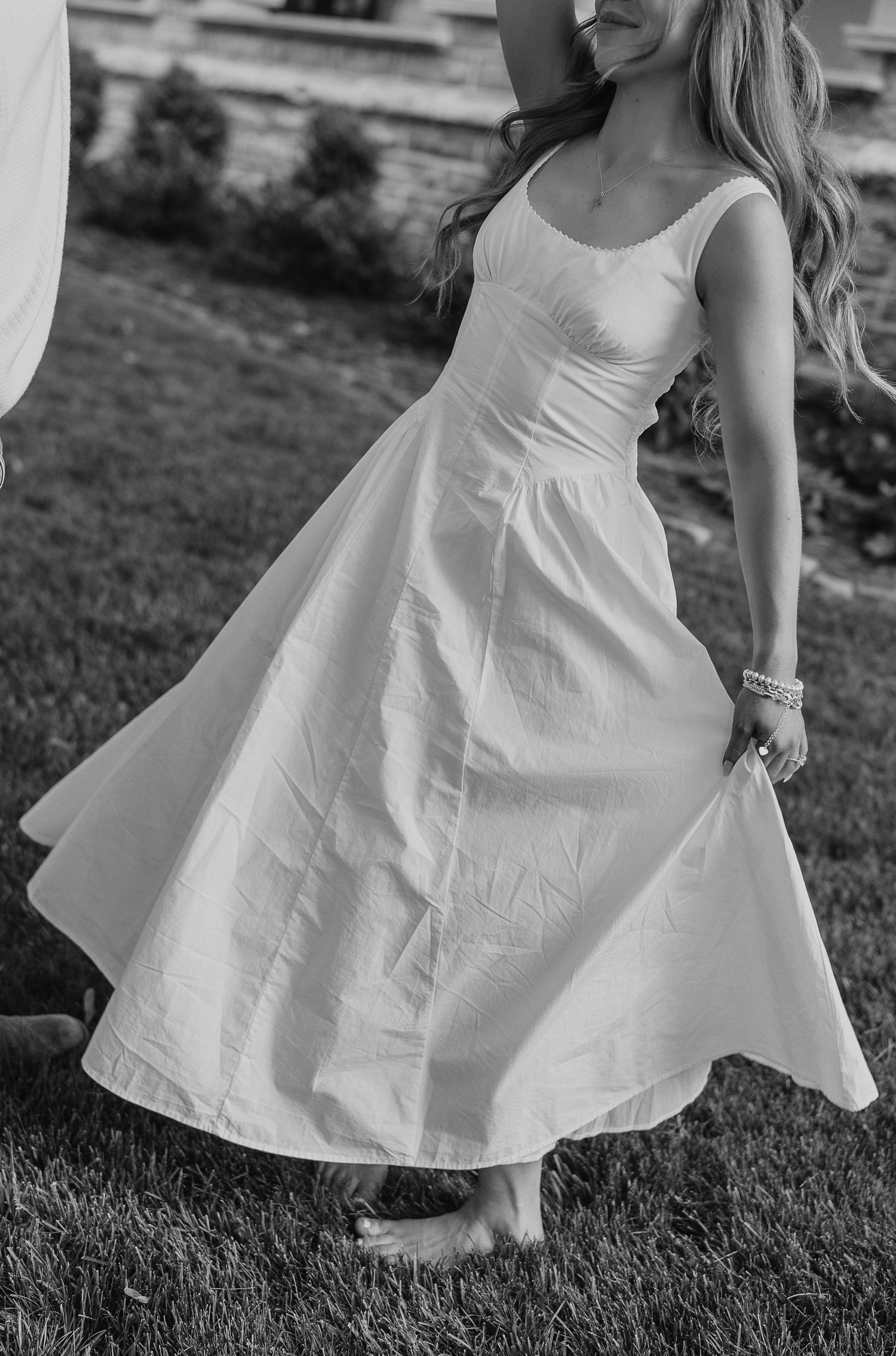 Woman in flowing white midi dress twirling barefoot on grass in black and white photo.