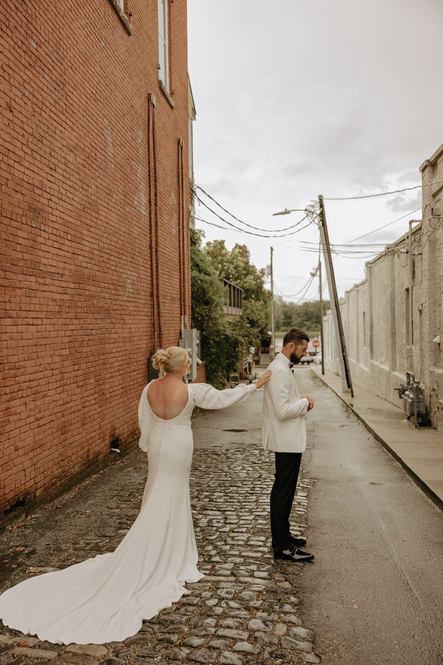A couple in wedding attire shares a first look moment in a cobblestone alley between brick buildings.