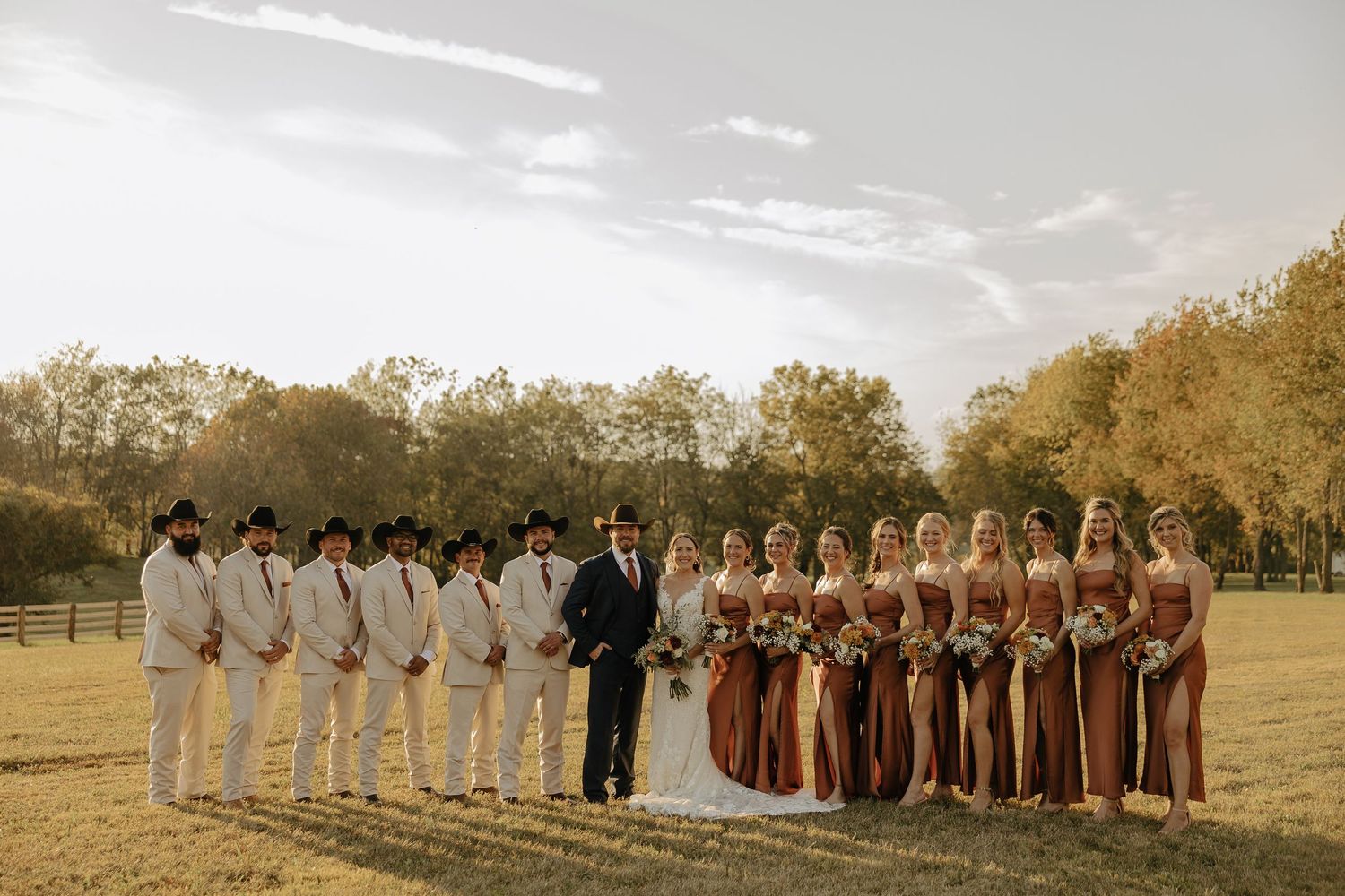 Large wedding party in white and brown attire posing together in a golden field at sunset.