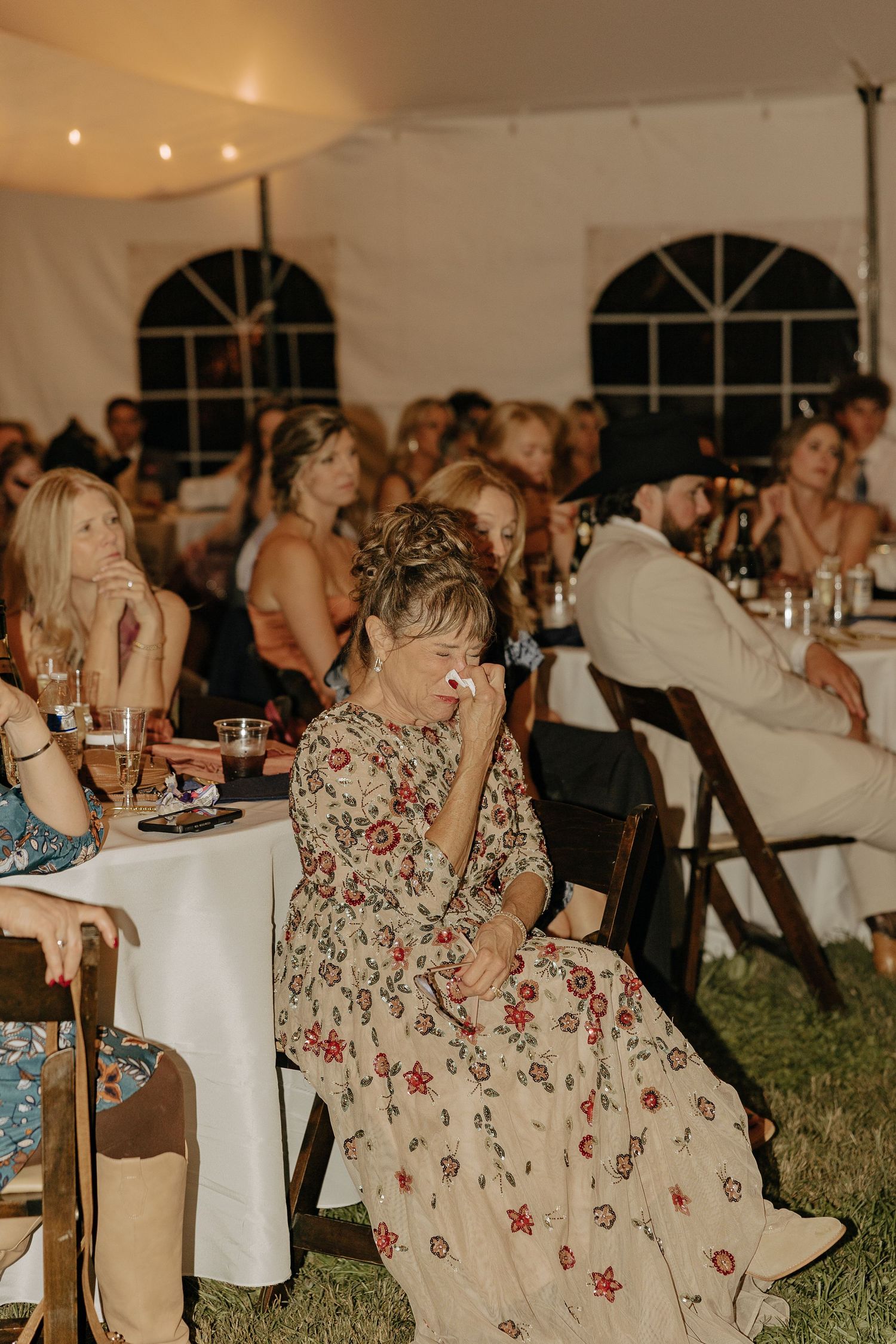 Wedding guests seated at tables in a tent venue wearing floral dresses and enjoying the celebration.
