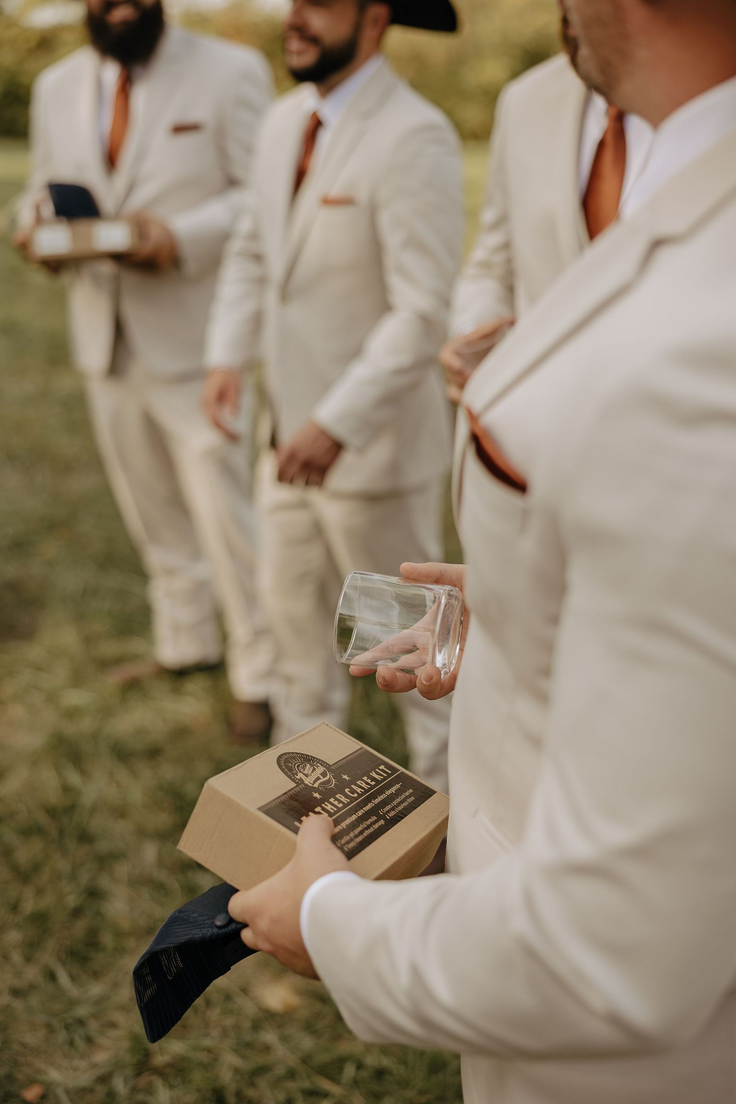 Groomsmen in light colored suits hold brown leather wallets during an outdoor wedding ceremony.