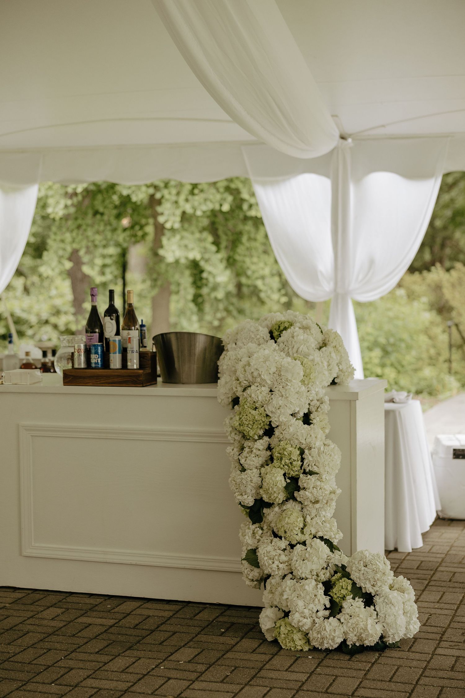 Elegant white outdoor wedding bar decorated with cascading white hydrangea florals under a draped tent.