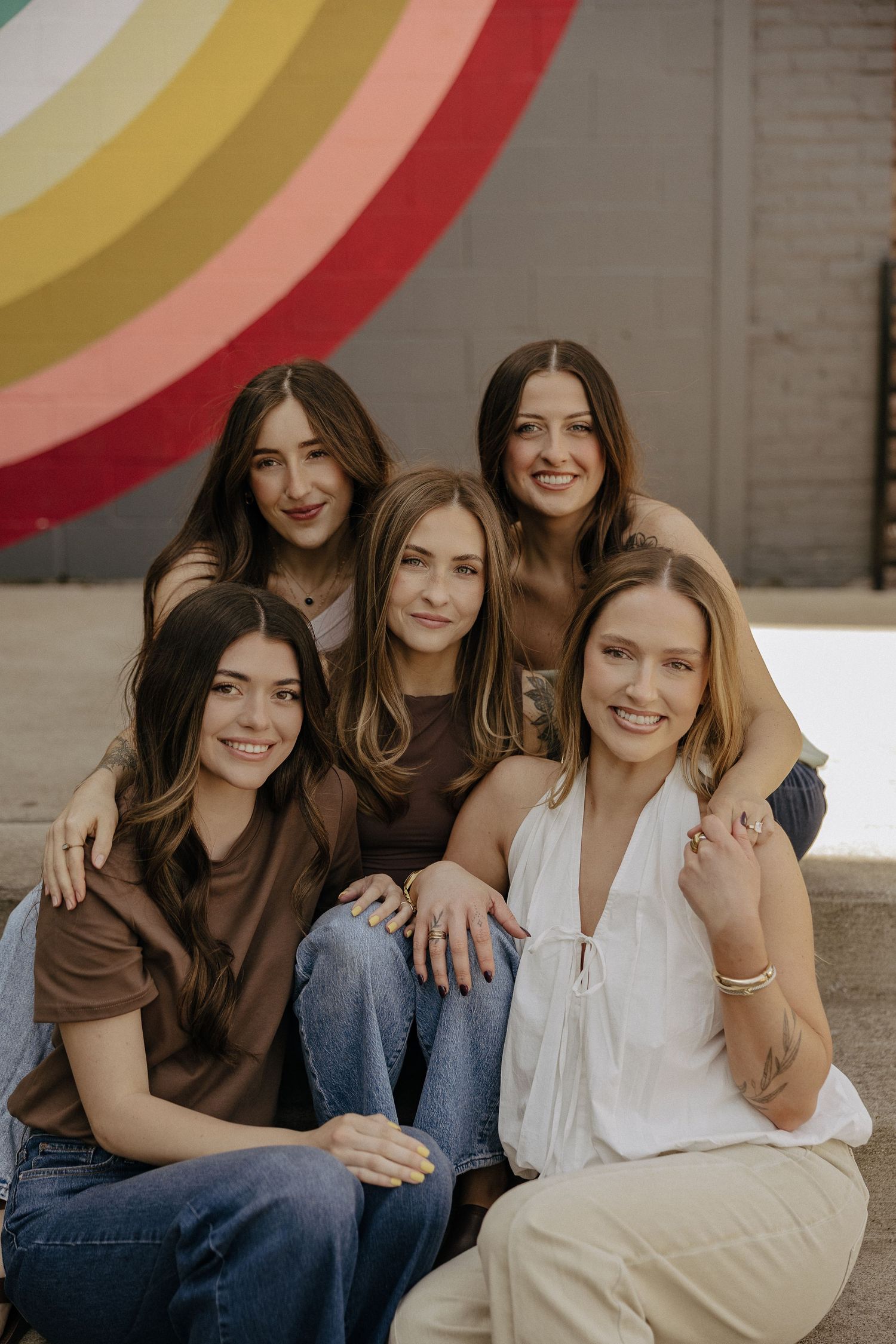 Five young women pose together smiling in front of a colorful rainbow mural outdoors.