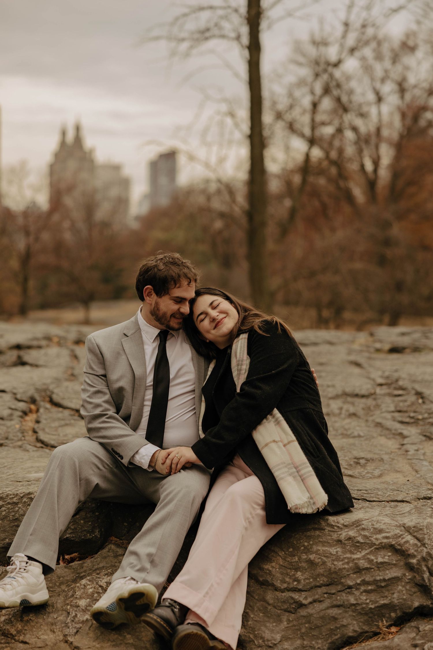 A couple sits intimately on rocky terrain during an autumn photoshoot with city buildings visible in background.
