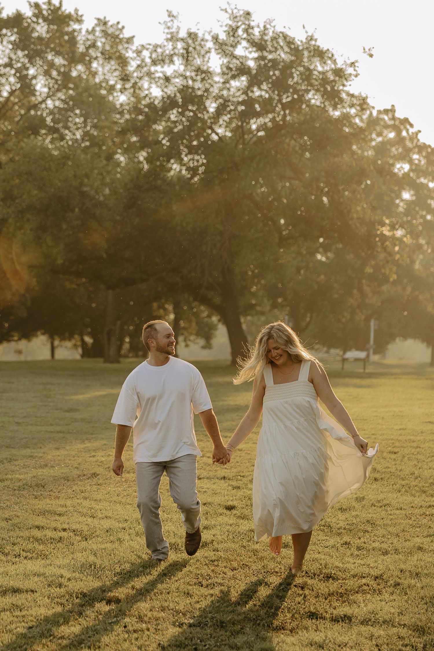 A couple holds hands while running through a sunlit grassy field at golden hour.