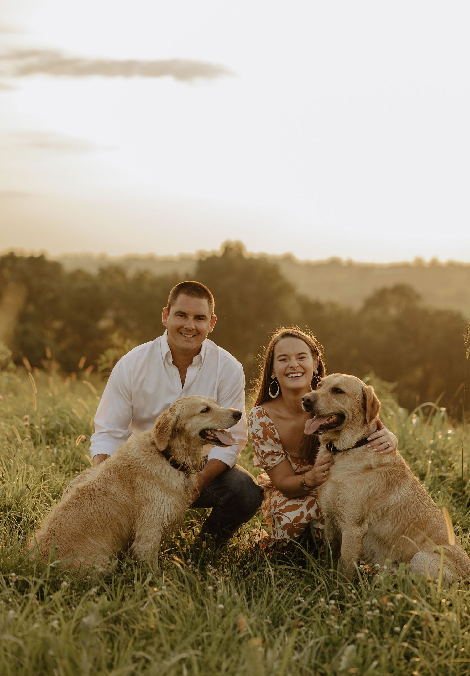 A couple sits with their three golden retriever dogs in a sunlit meadow during golden hour.
