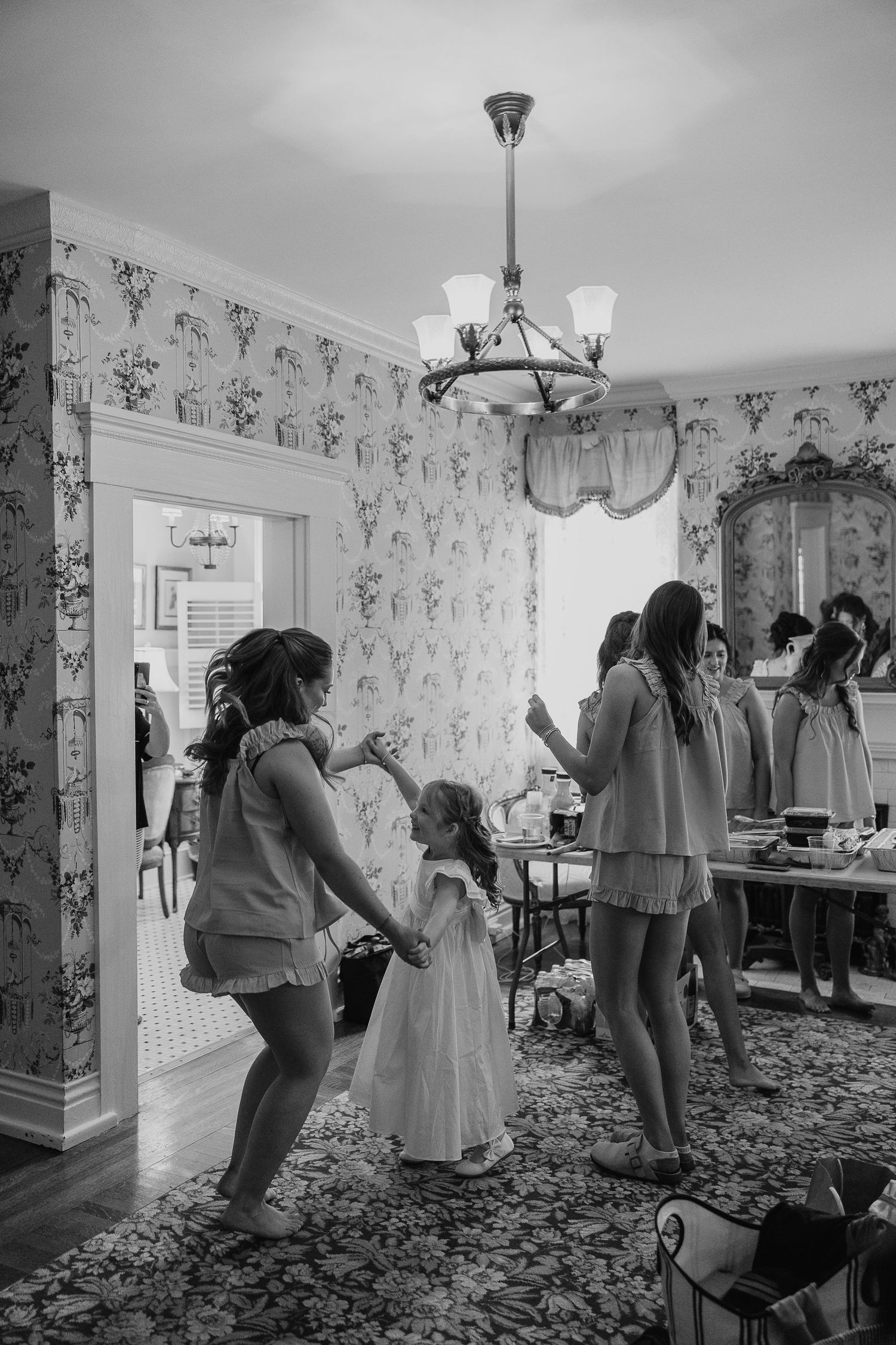 Bridesmaids in matching pajamas dance with flower girl in a vintage wallpapered bridal suite.