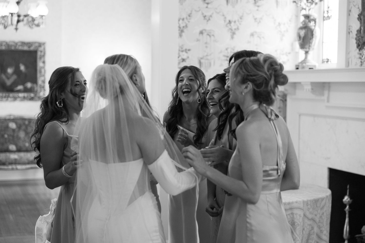 Bride in wedding dress and veil laughing with bridesmaids in elegant room, black and white photo.