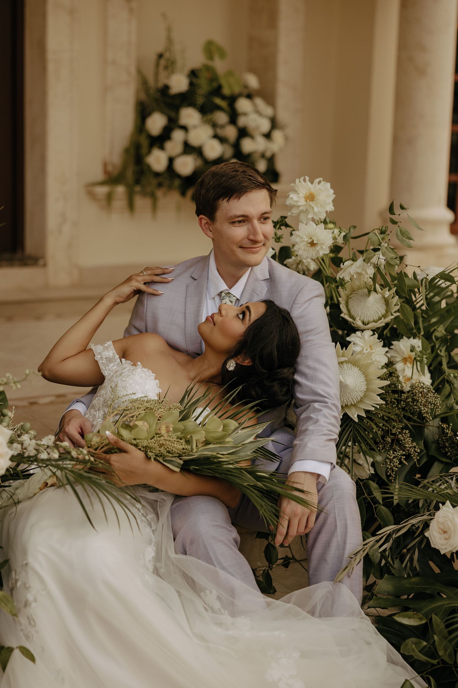A romantic wedding couple lounges together on a white couch surrounded by lush tropical greenery and white flowers.