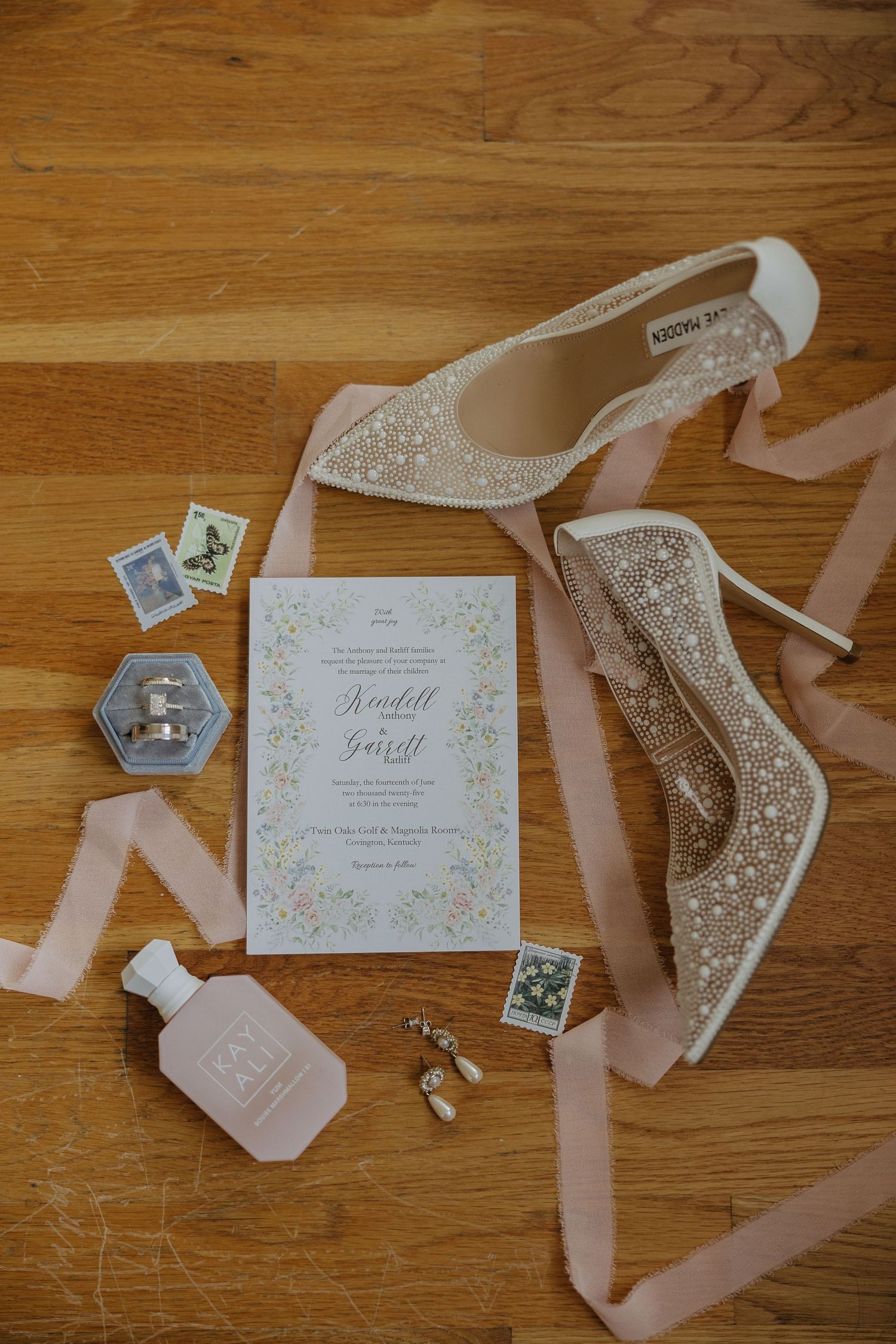 Wedding invitation and glittery bridal shoes arranged on wooden floor with pink ribbon and decorative elements.