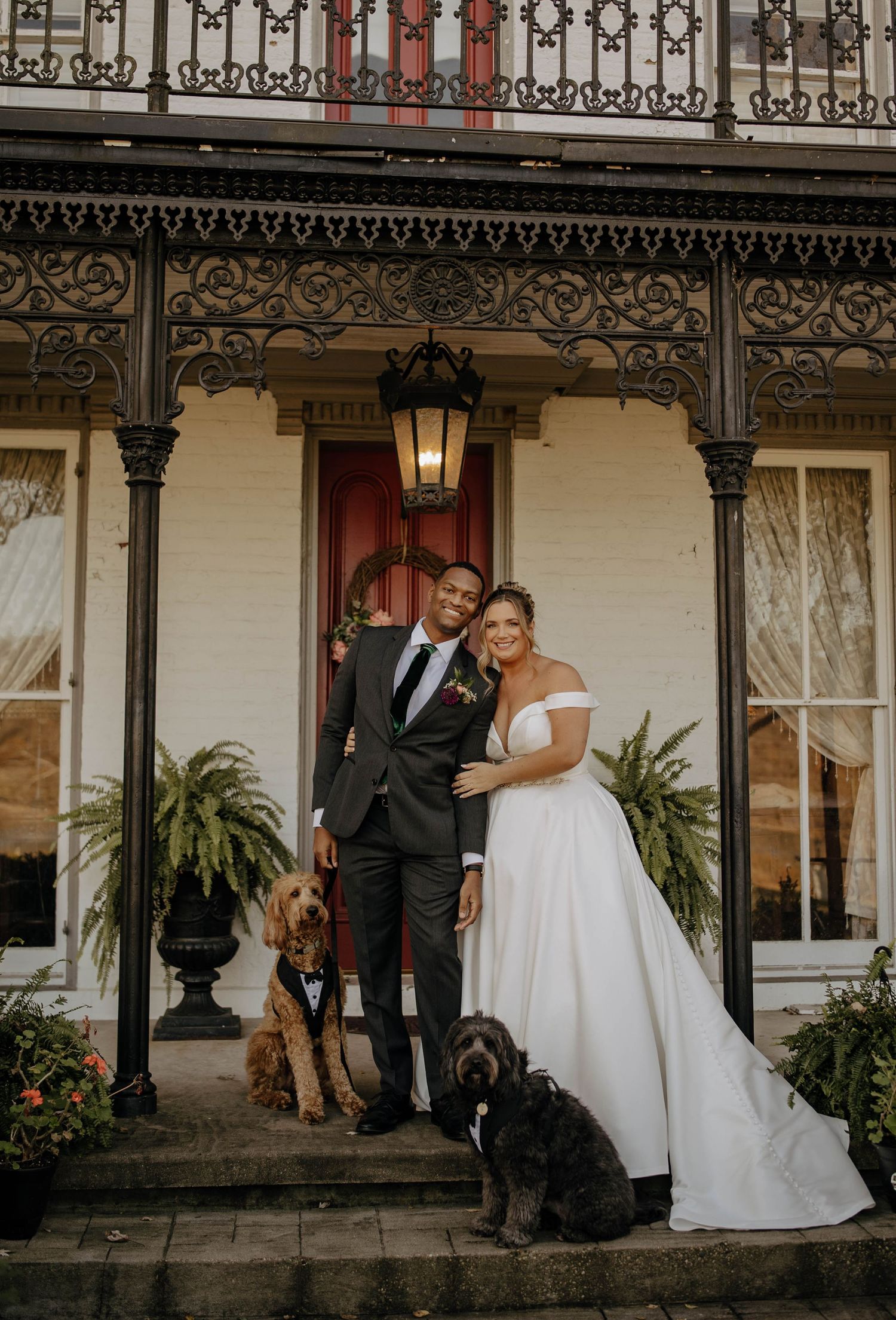 A couple in wedding attire poses on a Victorian porch with two dogs, under ornate ironwork and a vintage lantern.
