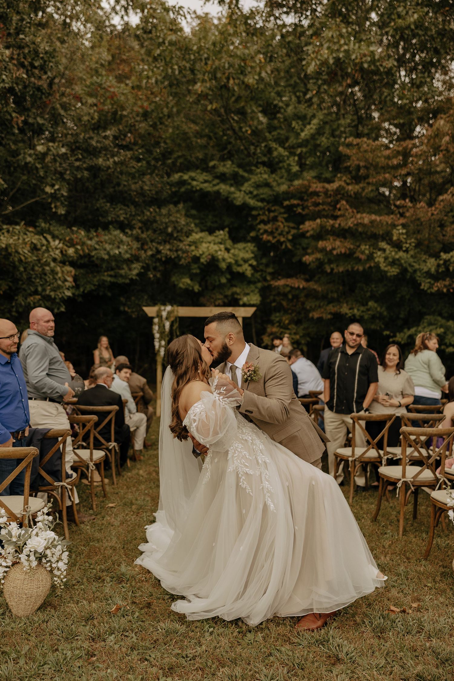 Wedding guests seated in wooden chairs watch a ceremony in a rustic outdoor setting.
