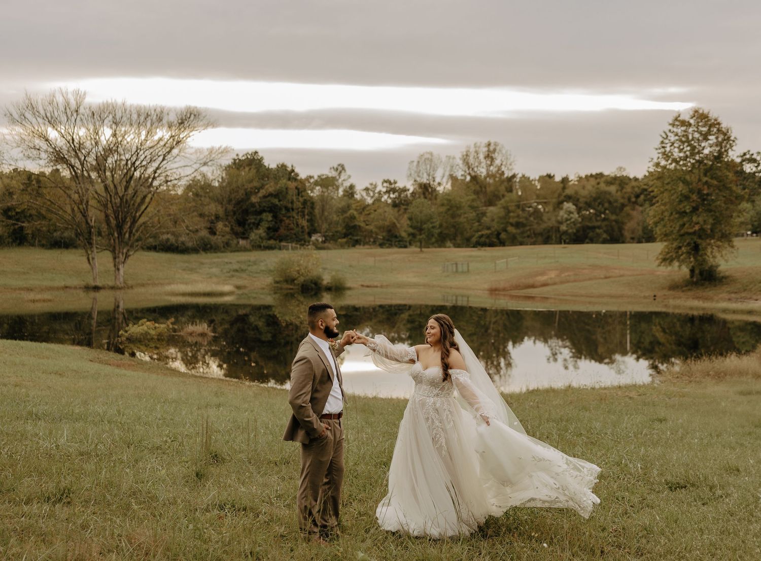 A couple walks through a scenic field with a lake in the background during their wedding photoshoot.