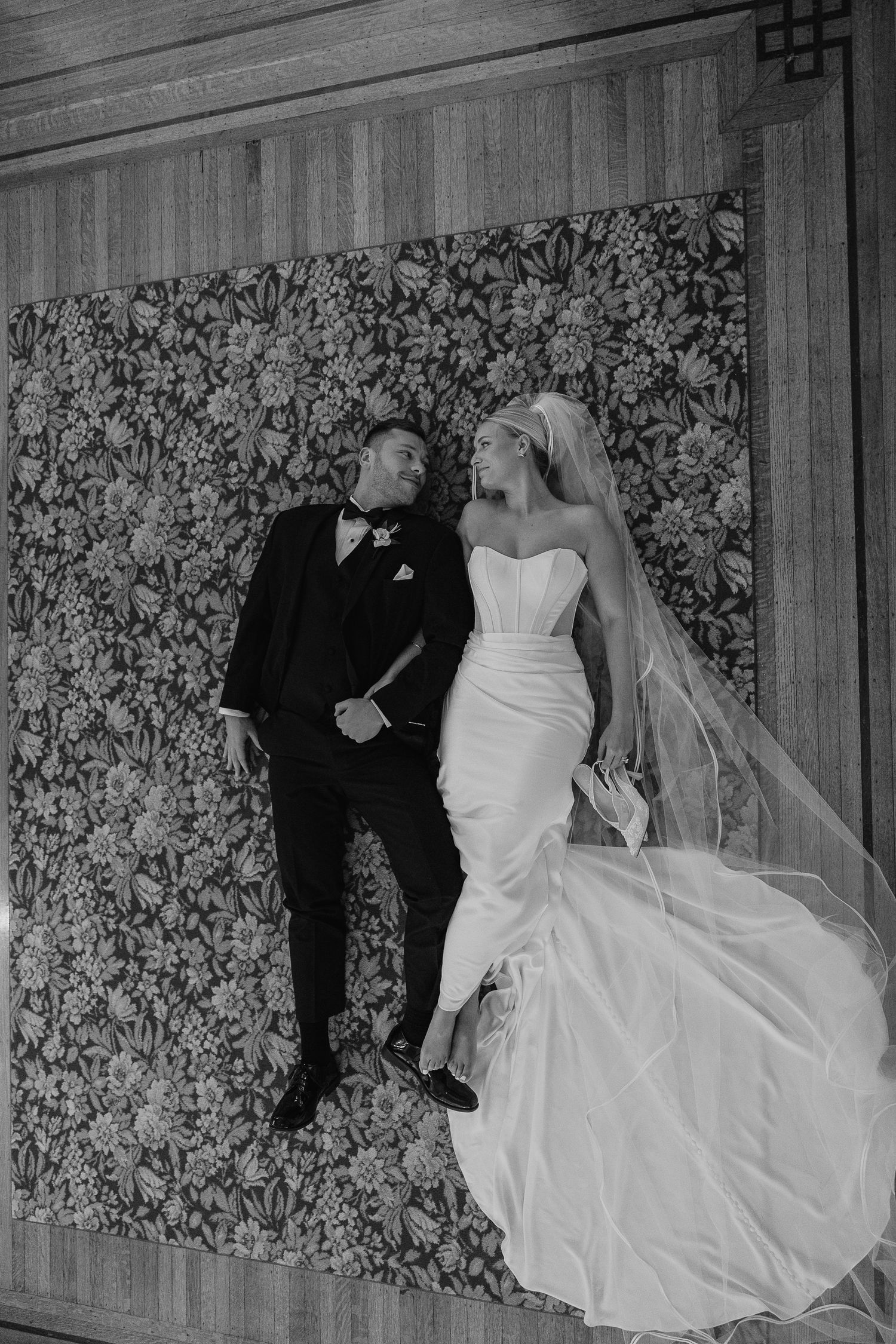 Black and white portrait of bride in strapless gown with veil and groom in tuxedo against floral wall.