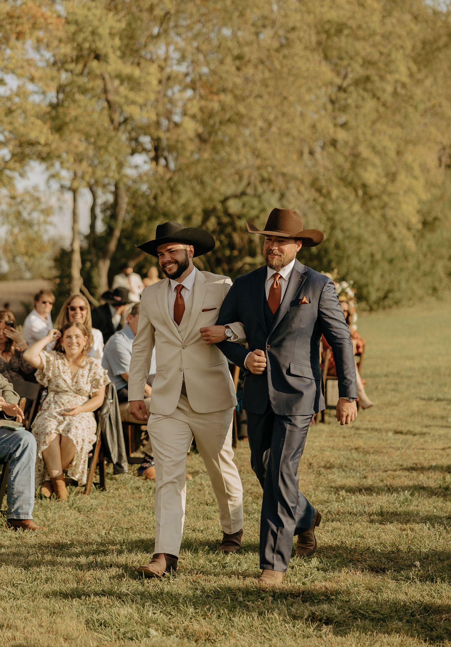 Two stylishly dressed men in suits and cowboy hats walk together on a grassy path during an outdoor event.