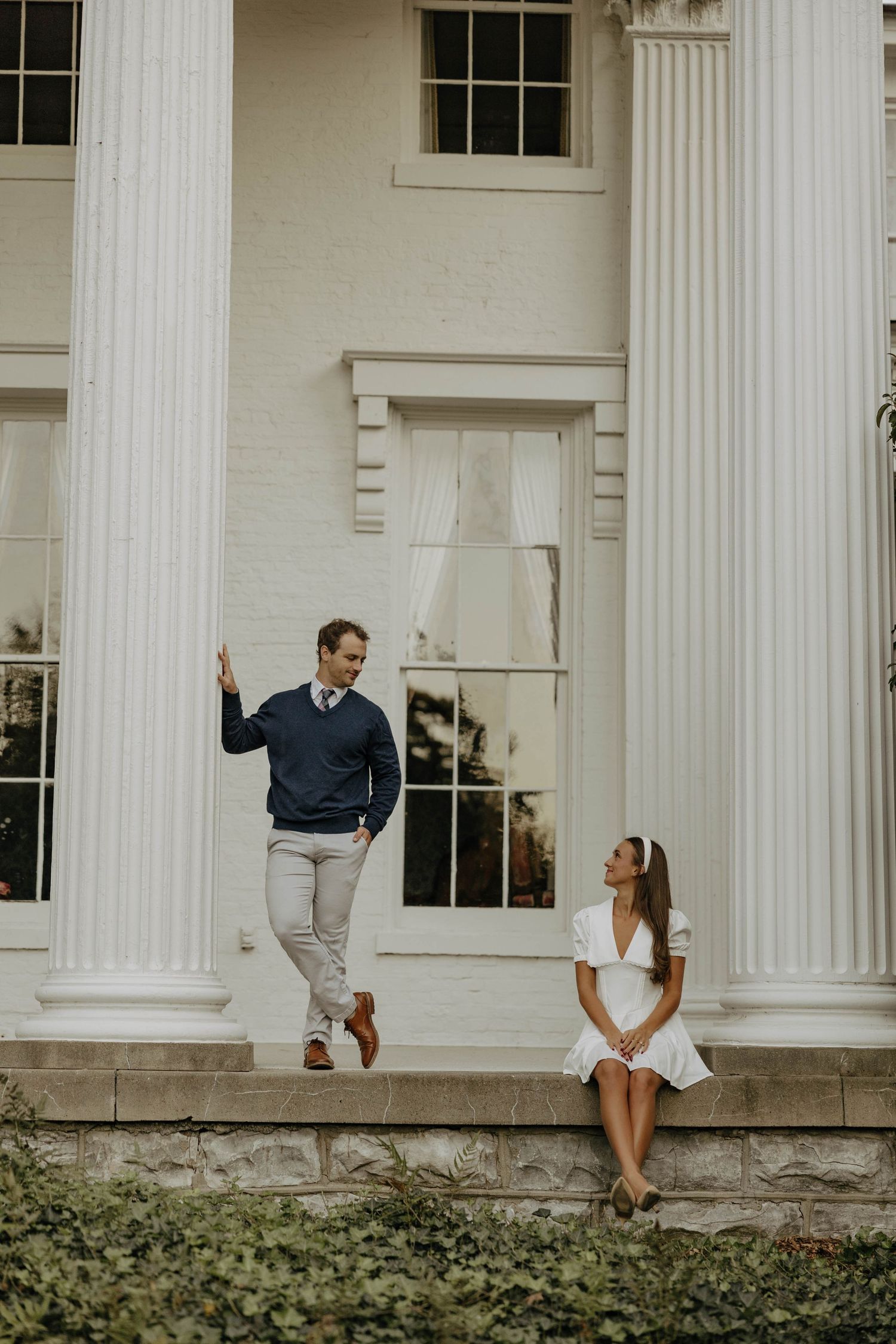 Couple poses playfully along white columned porch with landscaped grounds.