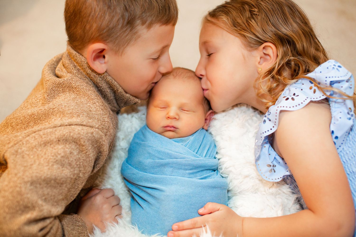 Two siblings kissing a sleeping newborn baby wrapped in a blue swaddle blanket.
