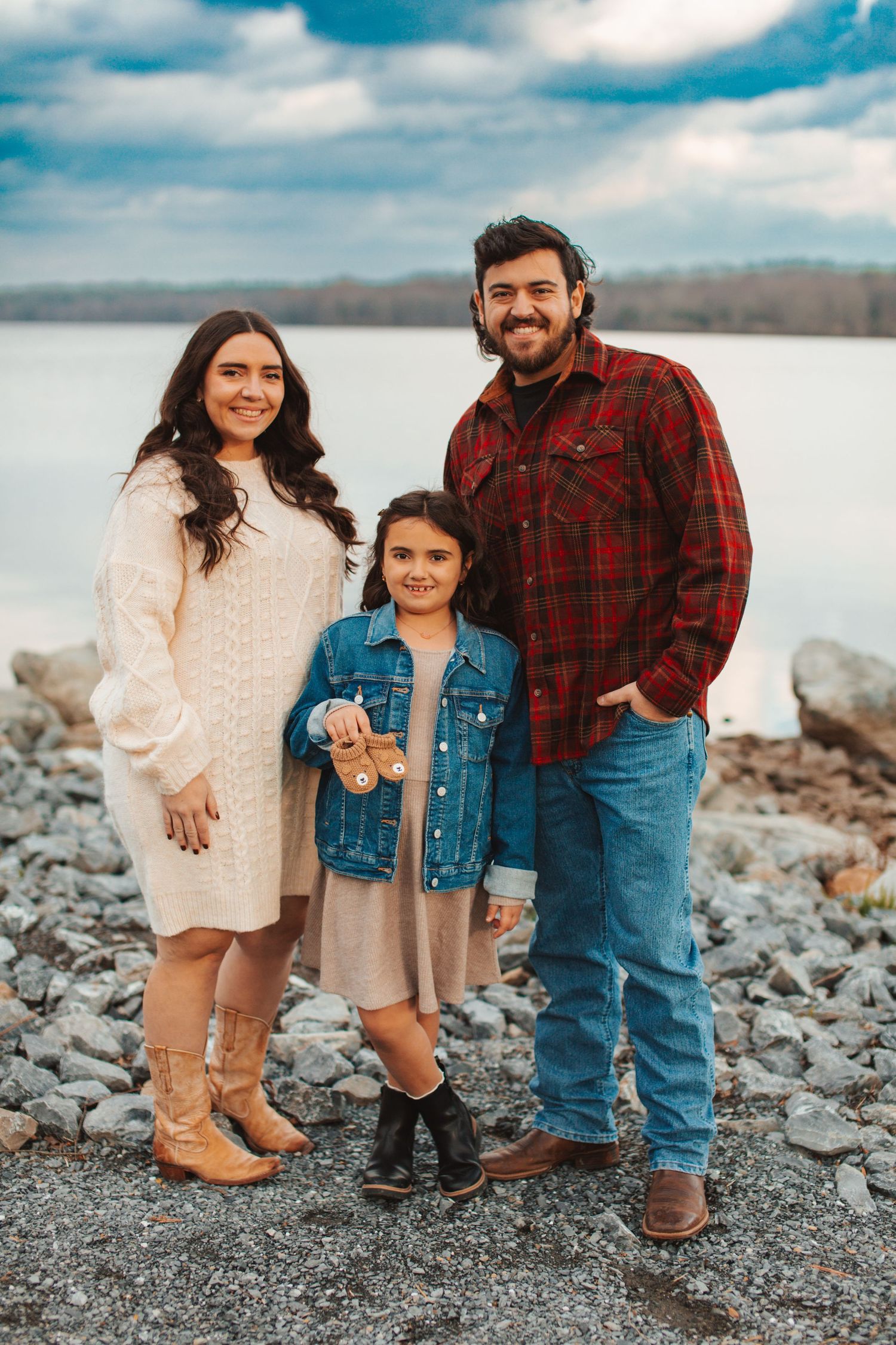 Family wearing coordinated neutral outfits poses together on rocky beach shoreline at sunset.