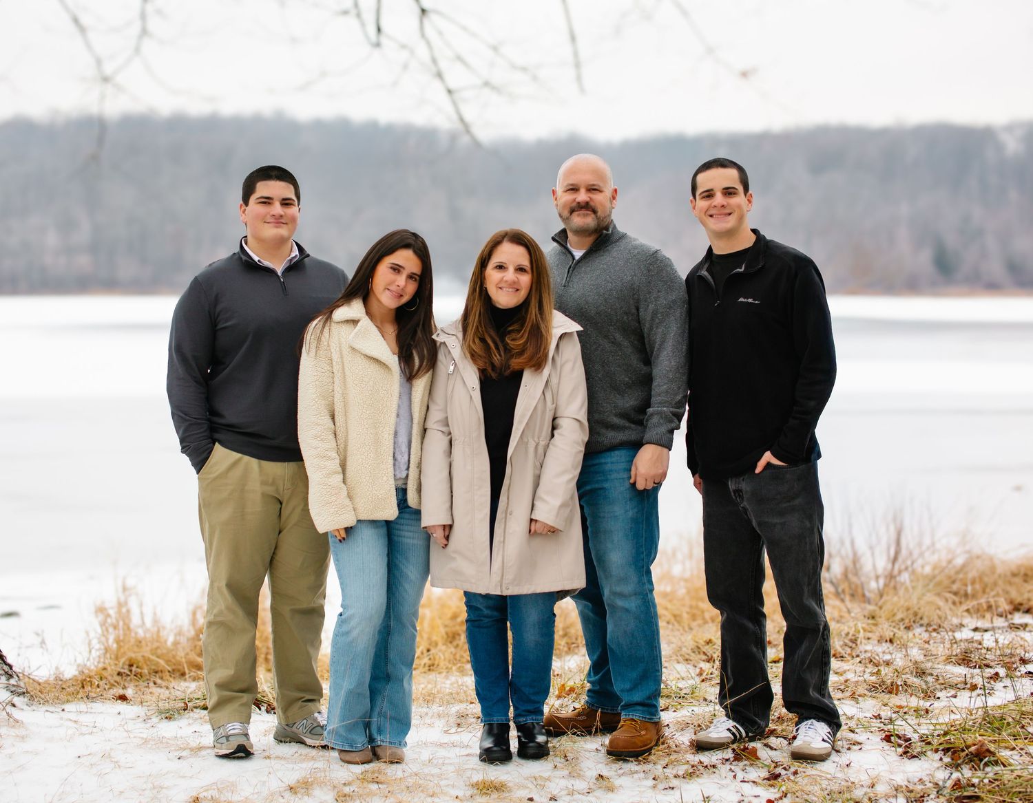 A group stands together outdoors in winter against a snowy lakeside backdrop wearing casual clothing.