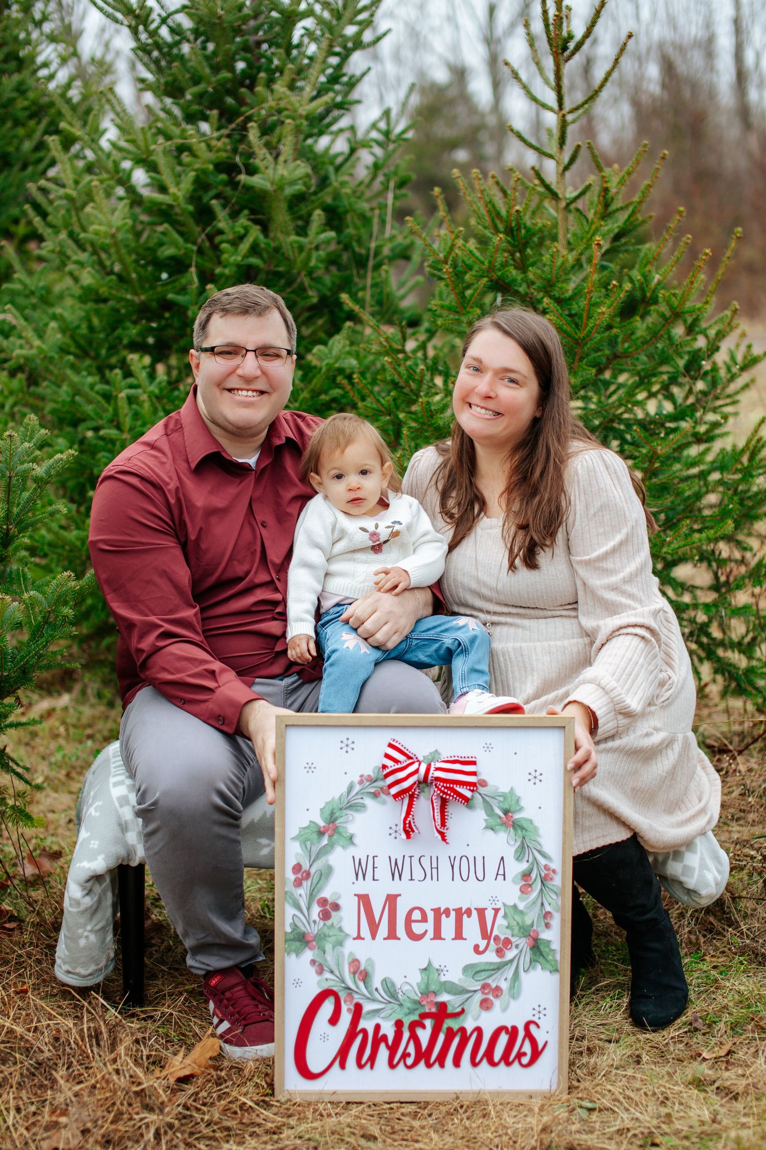 A family poses with festive Christmas signs among evergreen trees in a winter outdoor photo session.