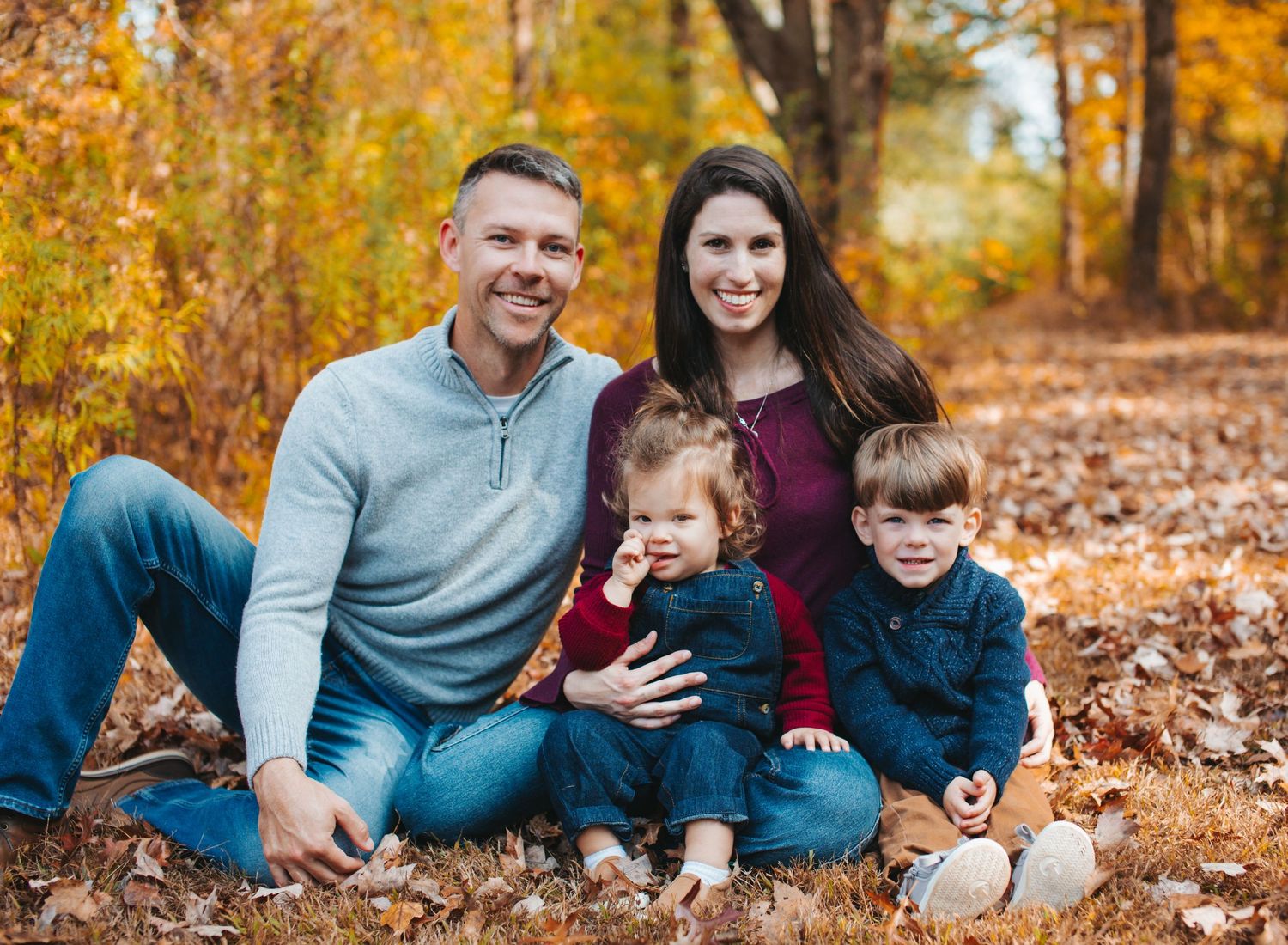 A family sits together on fallen autumn leaves with vibrant orange and yellow foliage in the background.