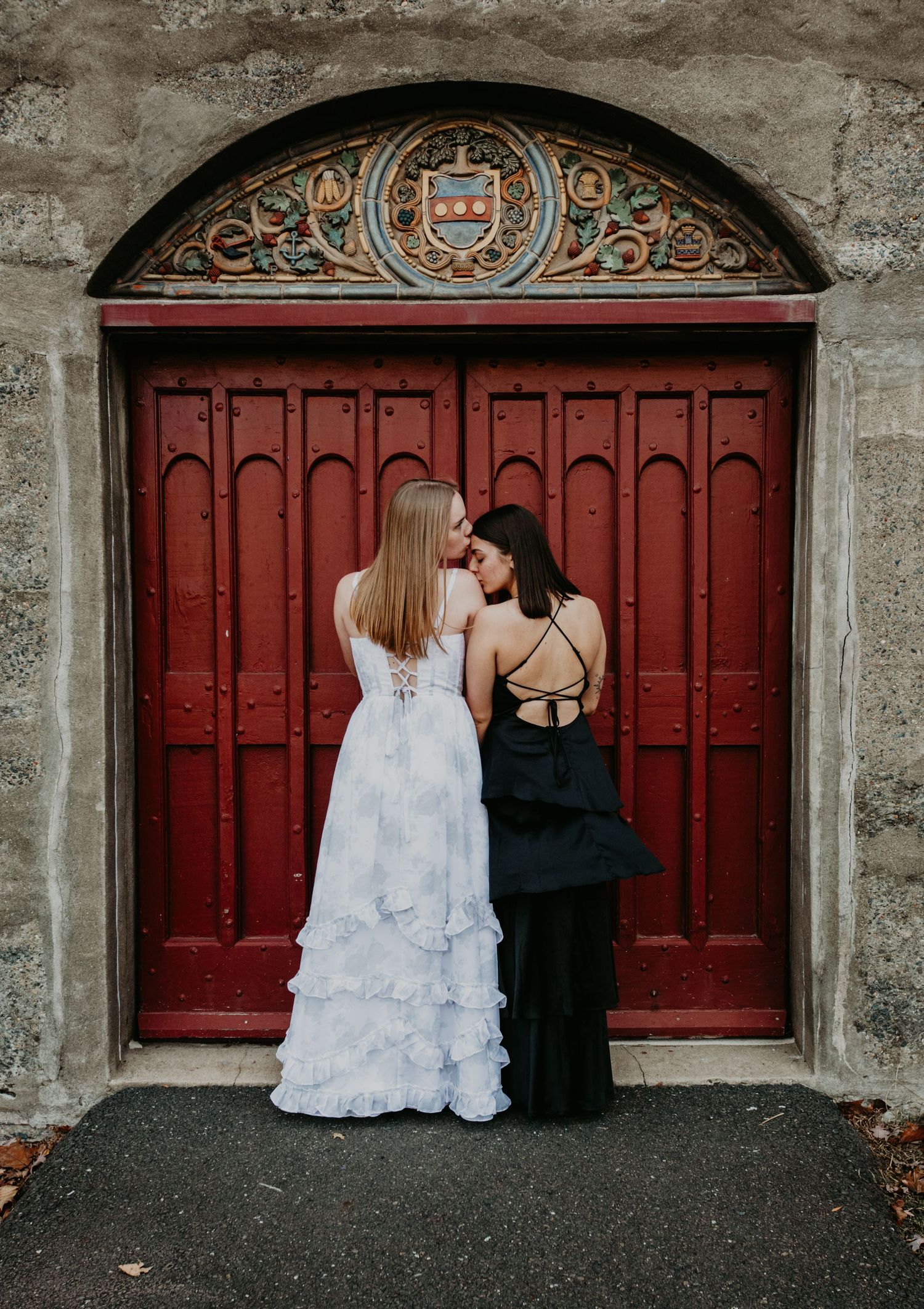 Two people in contrasting white and black dresses stand against a grand red door with ornate architectural details above.