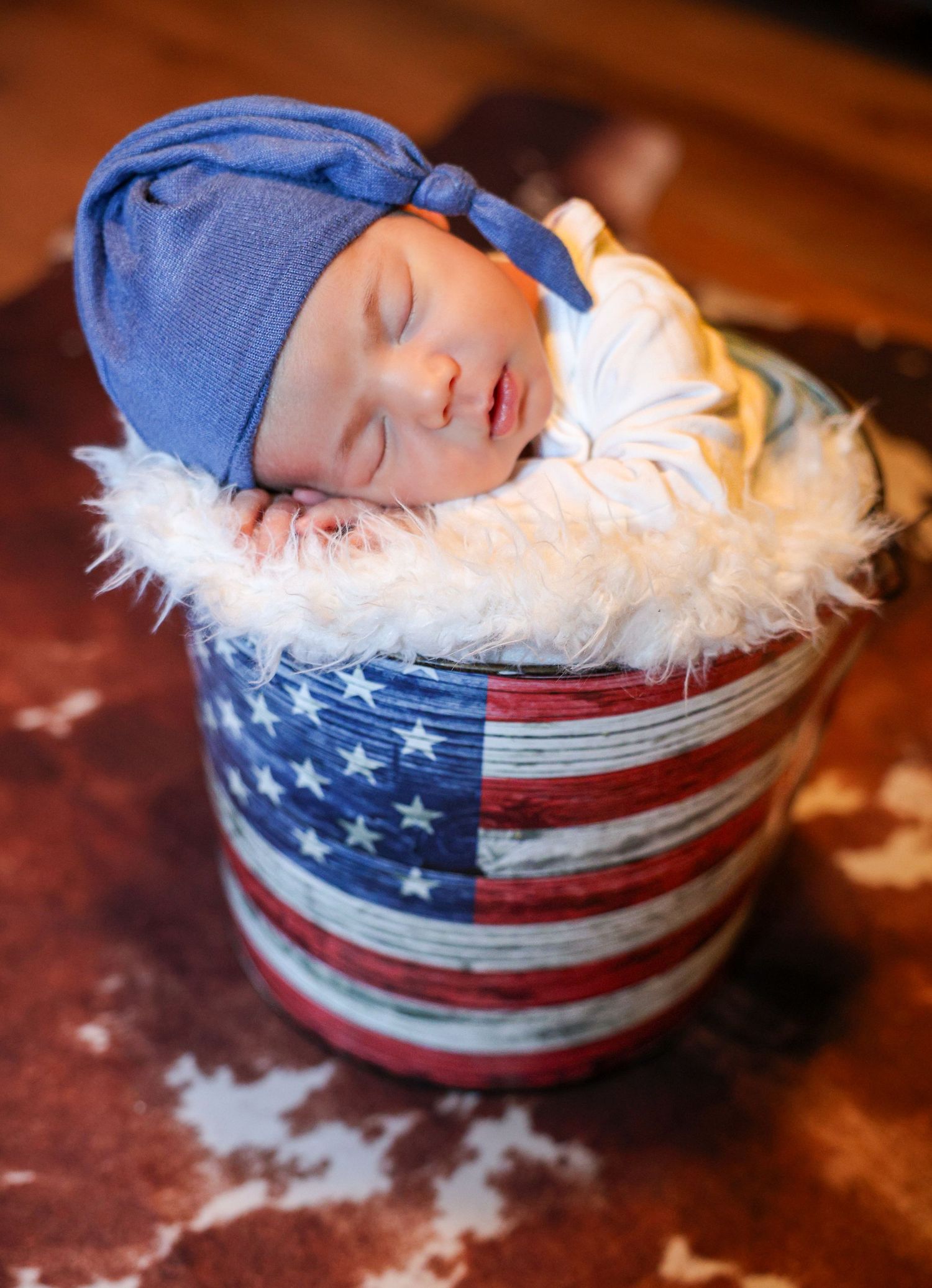 Sleeping newborn baby in blue knotted hat resting in American flag decorated bucket with white fur lining.