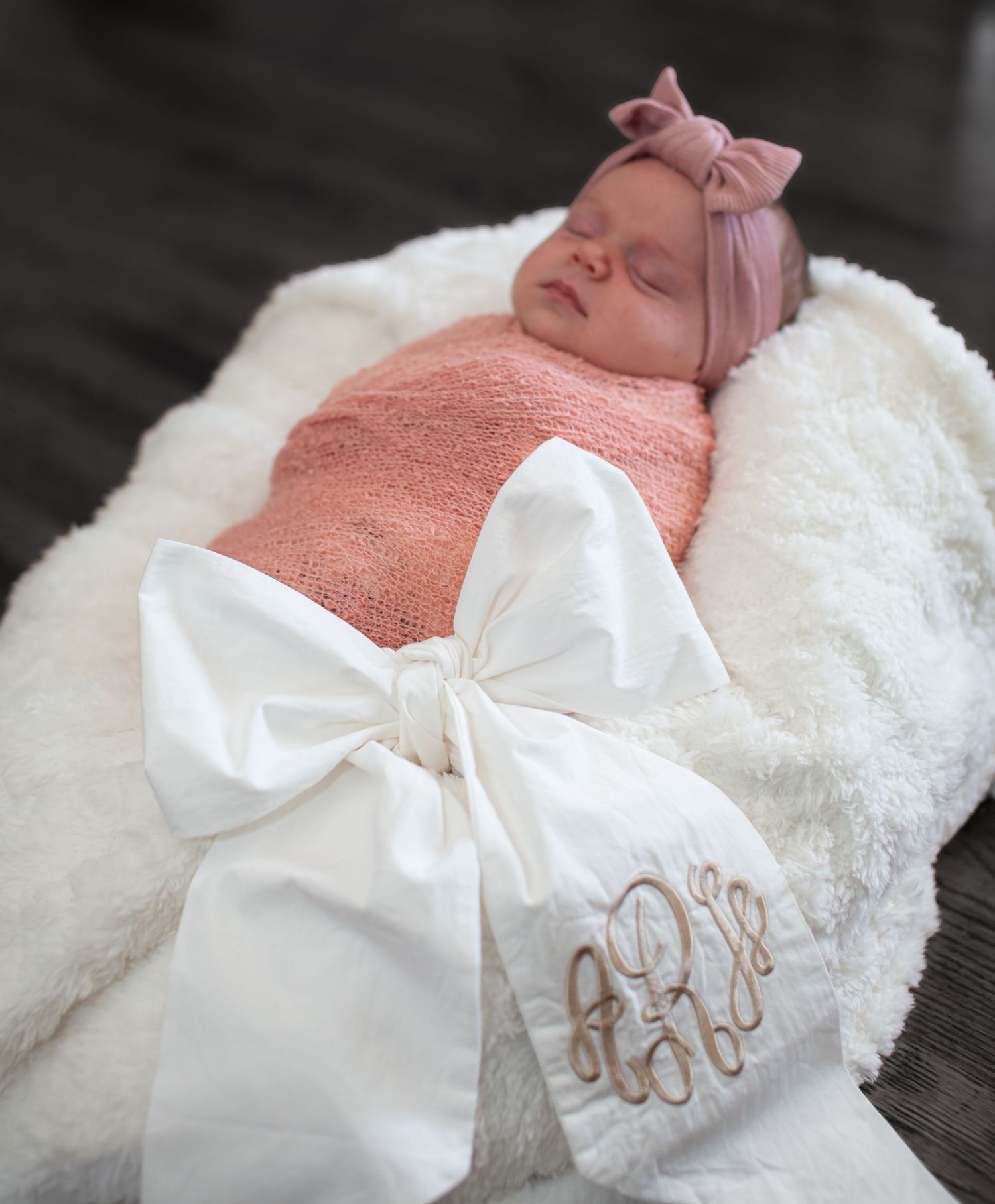 Newborn baby girl sleeping in white fur wrap with monogrammed bow, wearing pink knit swaddle and blush headband.