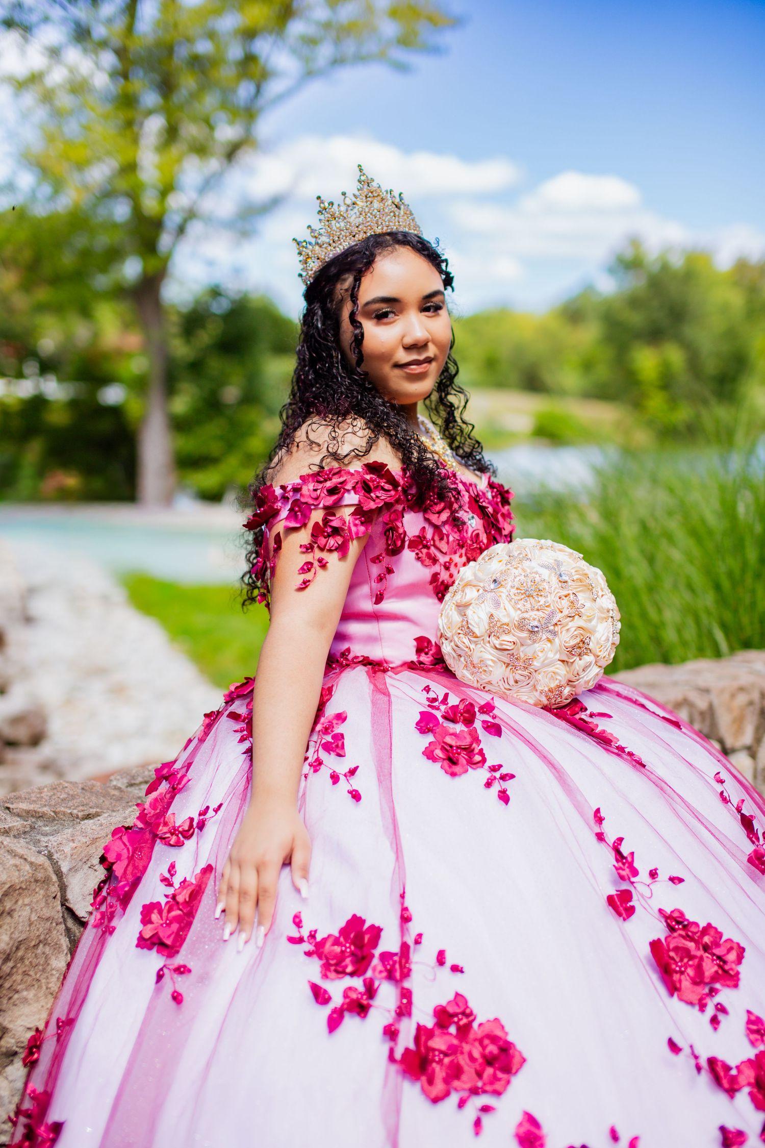 A beautiful quinceañera dress in white and pink with floral appliques photographed outdoors near a lake.