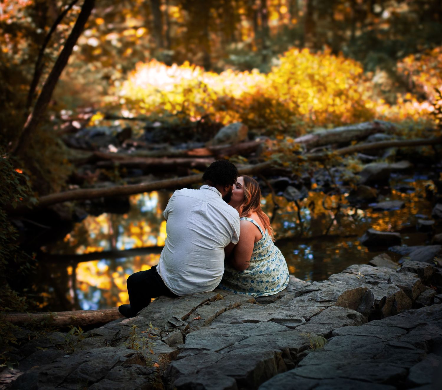 A couple sits together on rocky riverbank during golden sunset hour, creating a romantic silhouette against autumn foliage.