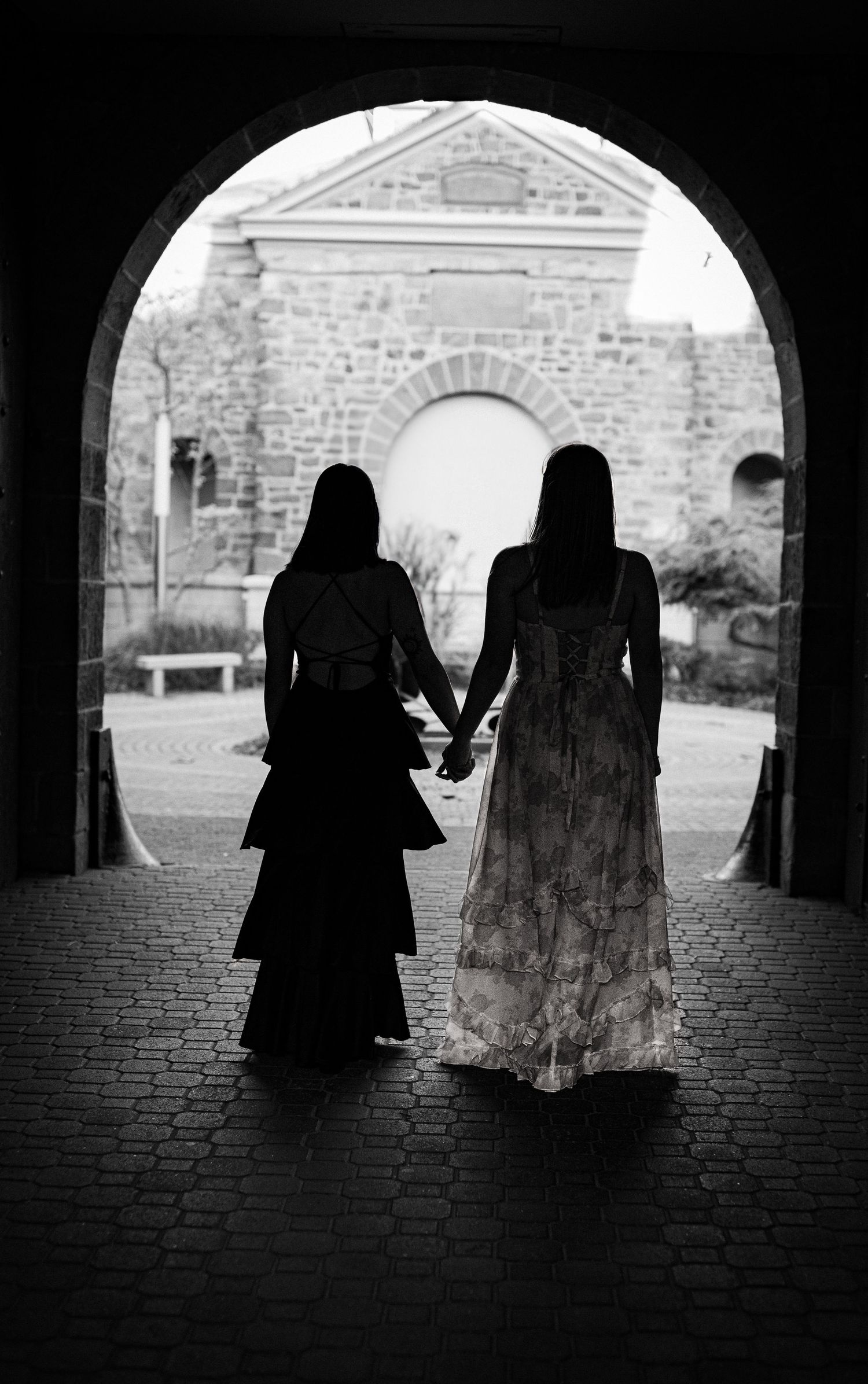 Silhouettes of two people in dresses stand under a stone archway in dramatic black and white.